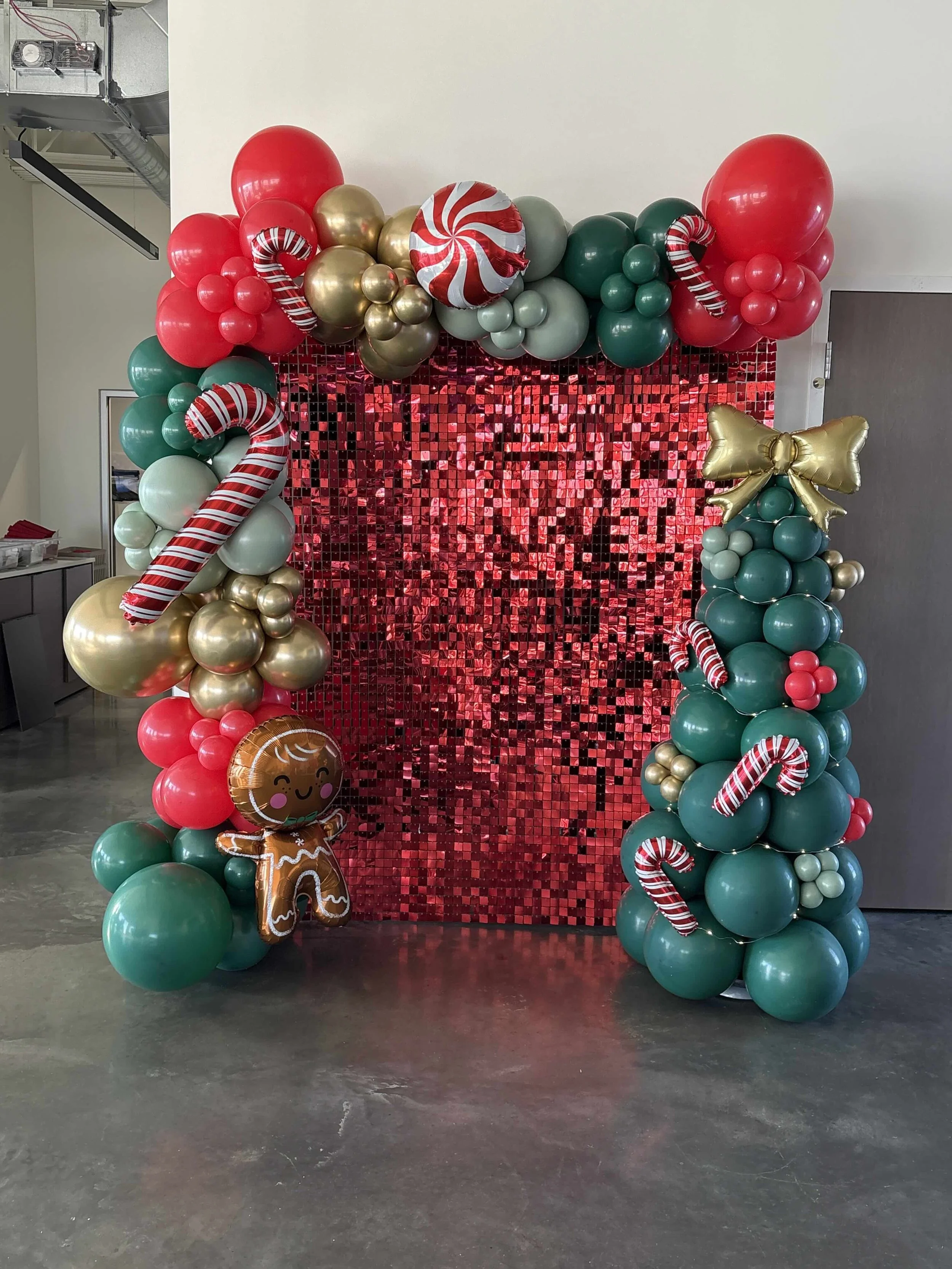 Holiday balloon display with red, green, gold, and striped balloons, candy cane and gingerbread themed balloons, and a shiny red sequin backdrop.