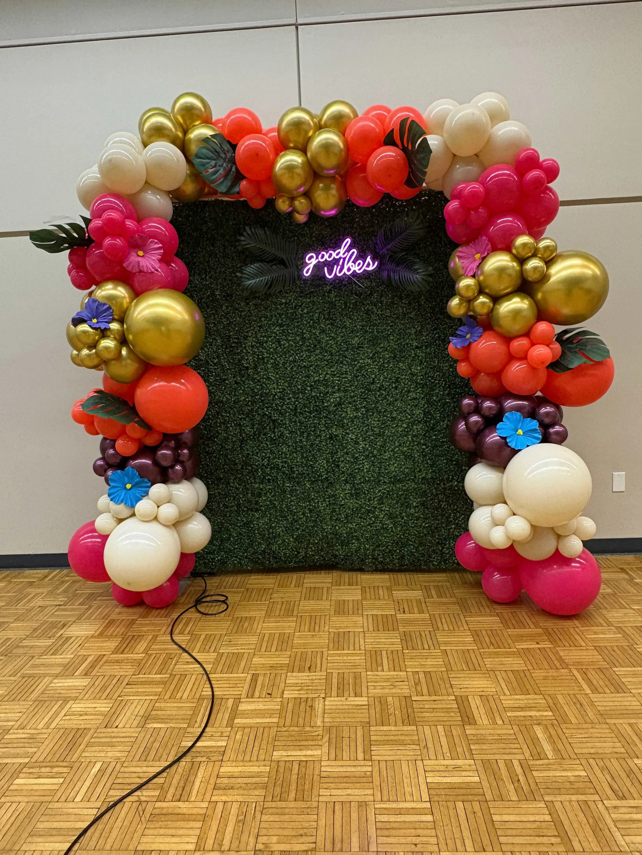 Decorative balloon arch with pink, white, gold, purple, and coral balloons surrounding a greenery backdrop with neon sign that says 'good vibes' and some tropical leaves, on a wooden floor.