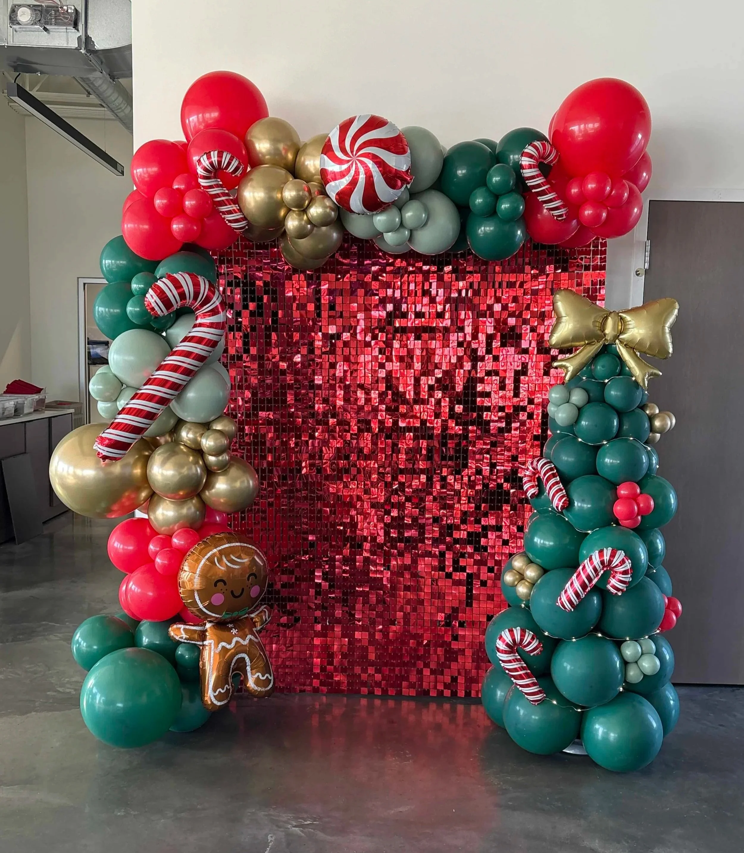 Festive balloon arch with red, gold, and green balloons, candy cane decorations, a gingerbread balloon, and a shiny red backdrop for holiday celebration.