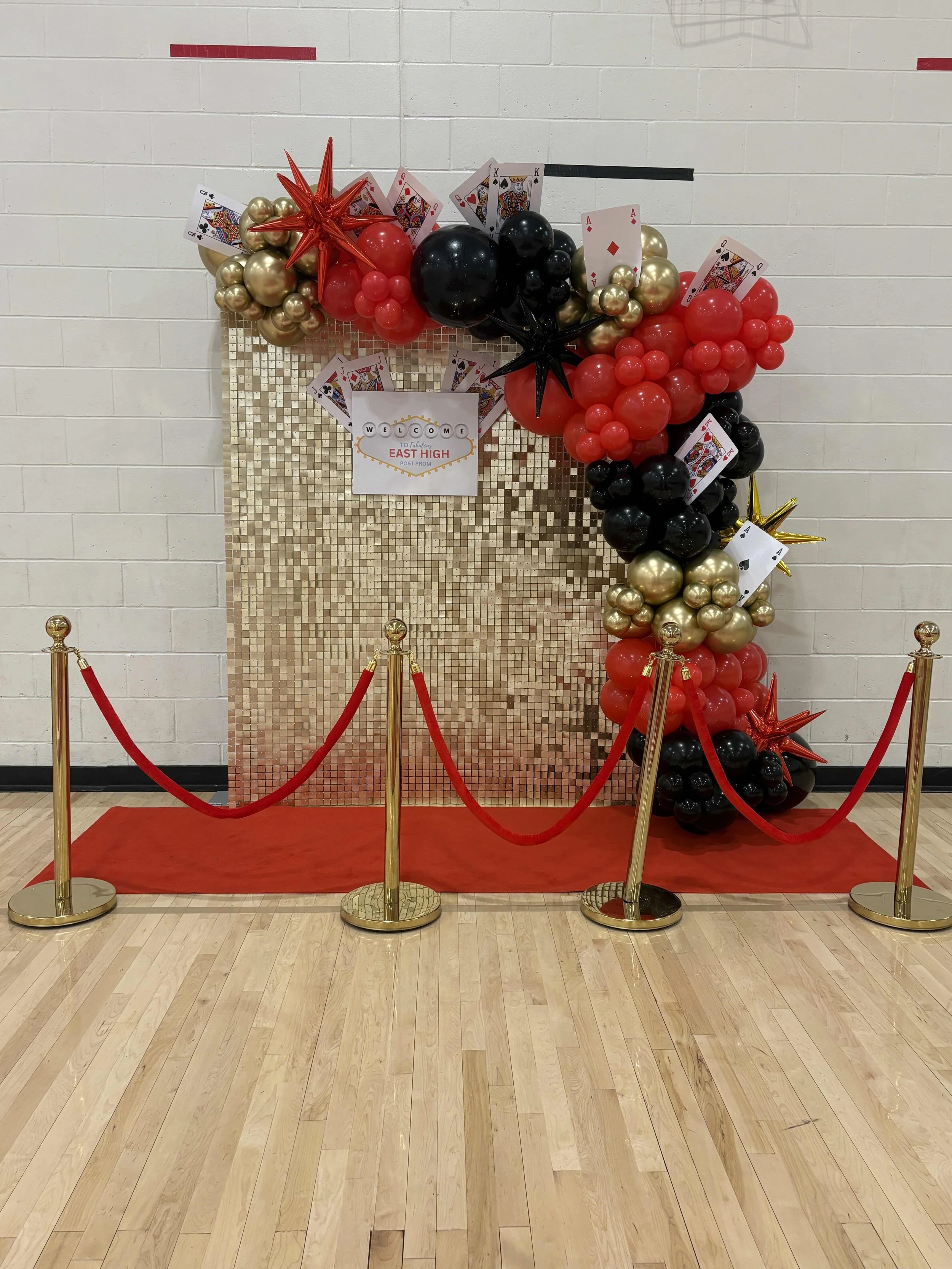 Decorative backdrop with balloons in black, red, and gold, playing cards, and a sign that says 'Welcome to East High Post Prom.' A red carpet with gold stanchions and red ropes is in front of the backdrop.