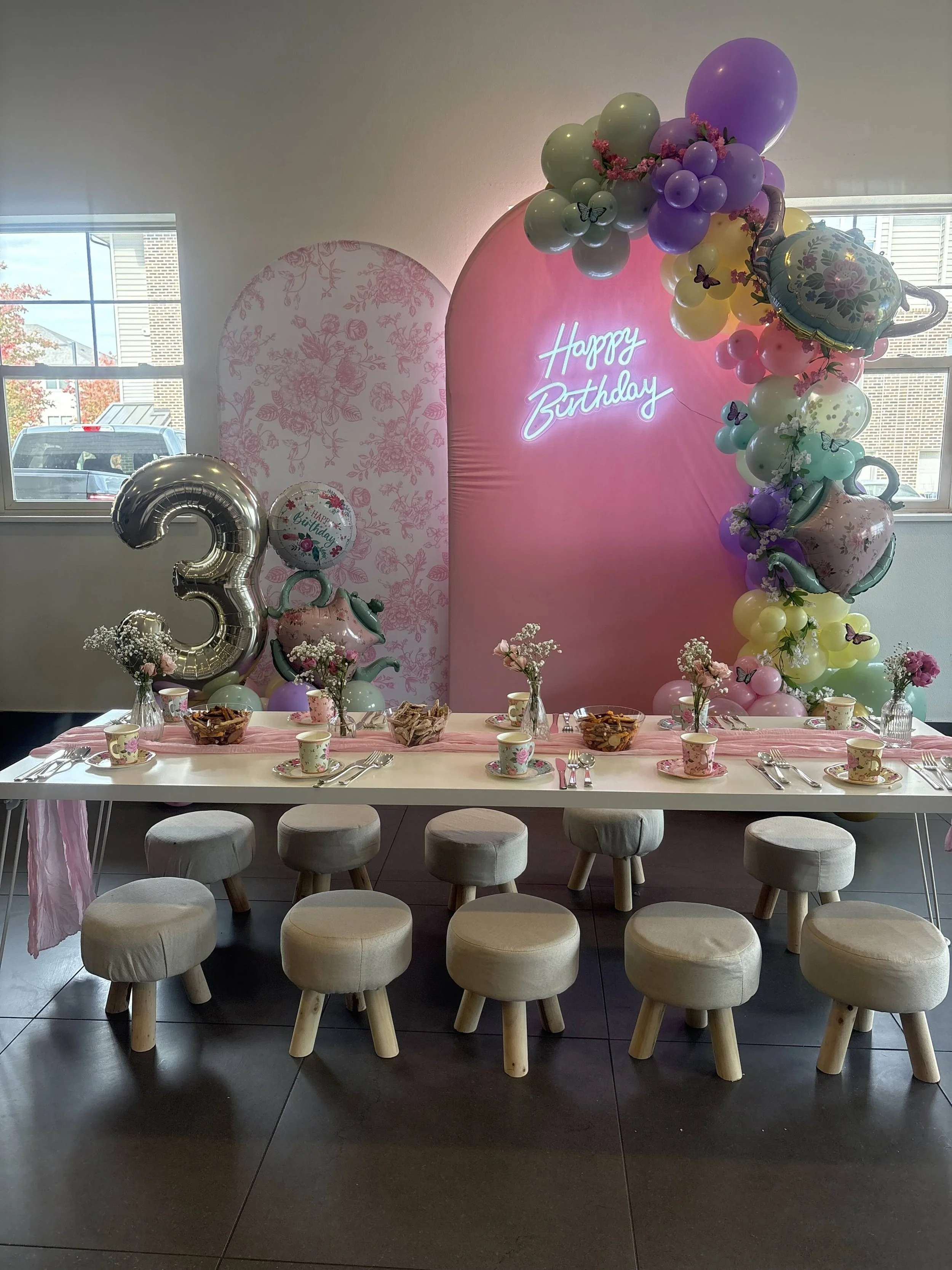 Decorated birthday party table with flower arrangements, balloons, and a 'Happy Birthday' neon sign backdrop.