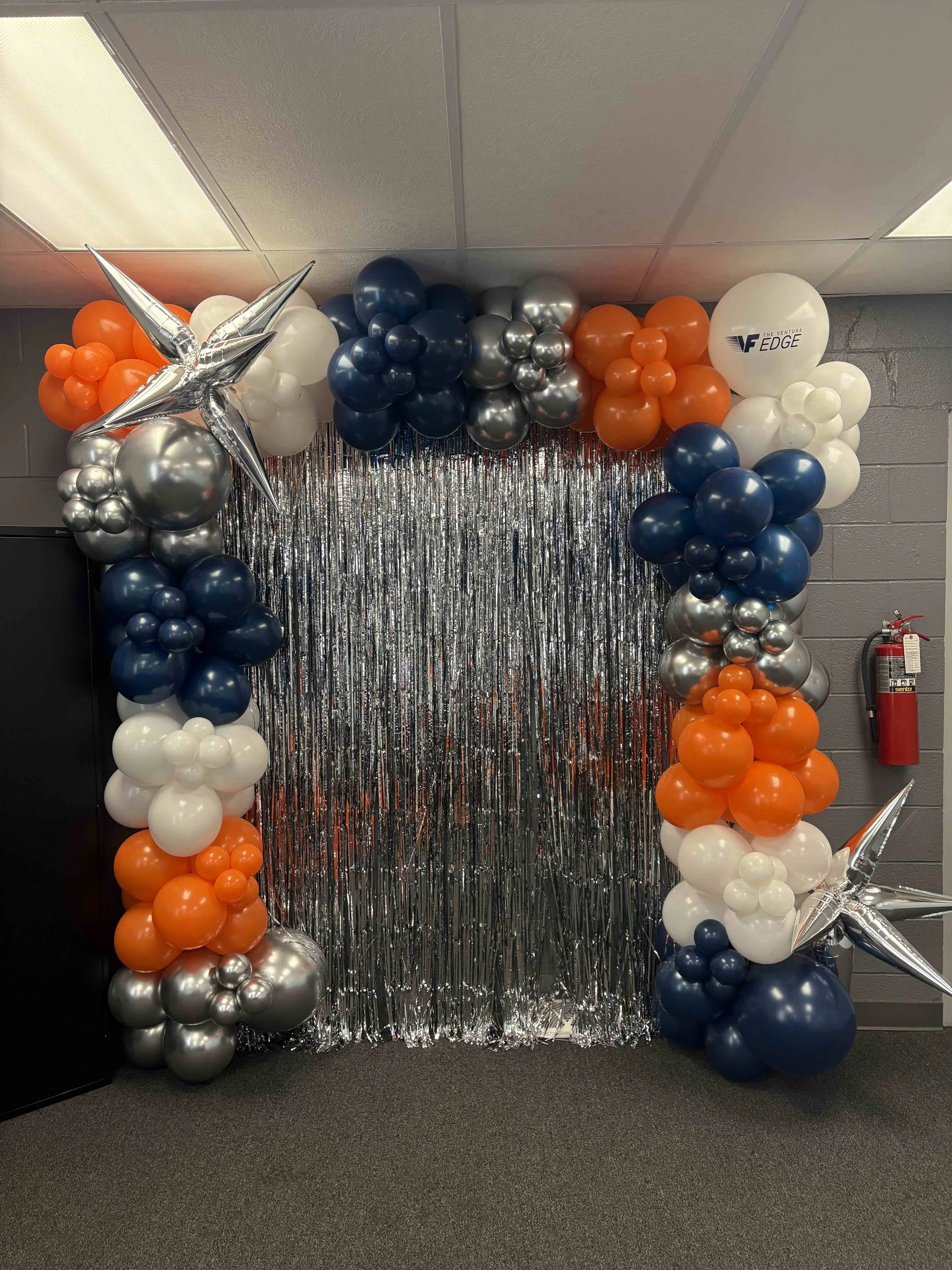 Balloon arch with orange, white, navy, and silver balloons, silver star-shaped balloons, and silver fringe backdrop.
