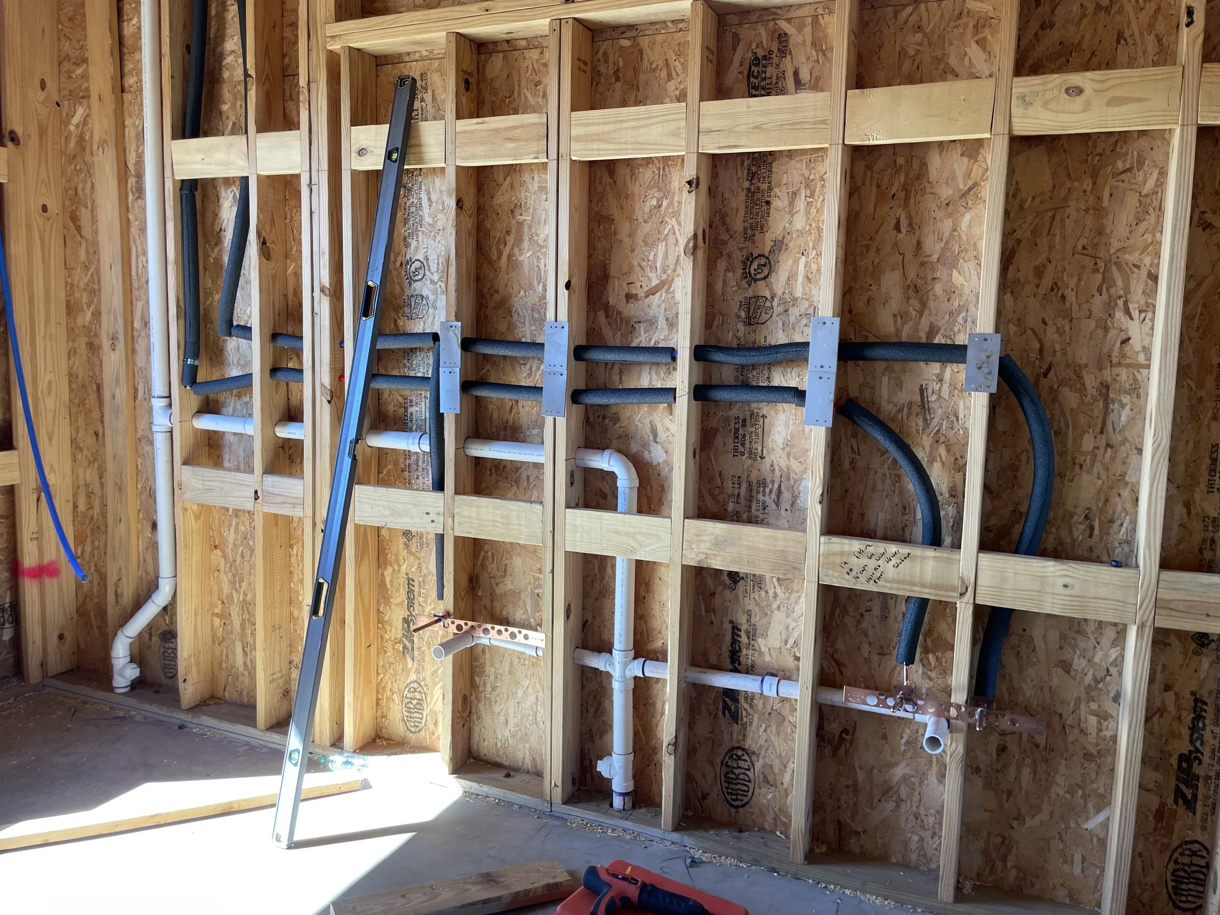 Interior wall with exposed wood framing, electrical and plumbing pipes, and hoses installed for future fixtures in a construction site.