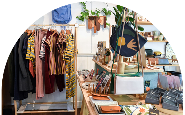 Display of clothing, handbags, and accessories in a boutique store with a clothing rack holding striped and solid-colored shirts and jackets, and a table with wallets, phone cases, and jewelry.