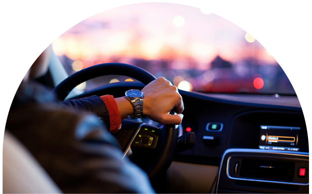 Close-up of a driver's hand on steering wheel inside a vehicle at sunset, with blurred dashboard and city lights outside