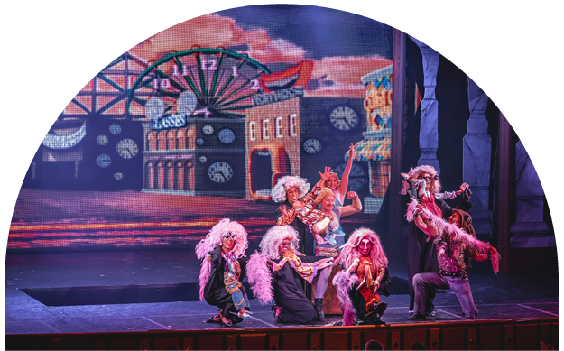Group of performers dressed in colorful costumes with wigs and makeup, performing a dance on stage with a carnival or amusement park backdrop featuring a Ferris wheel and a roller coaster.