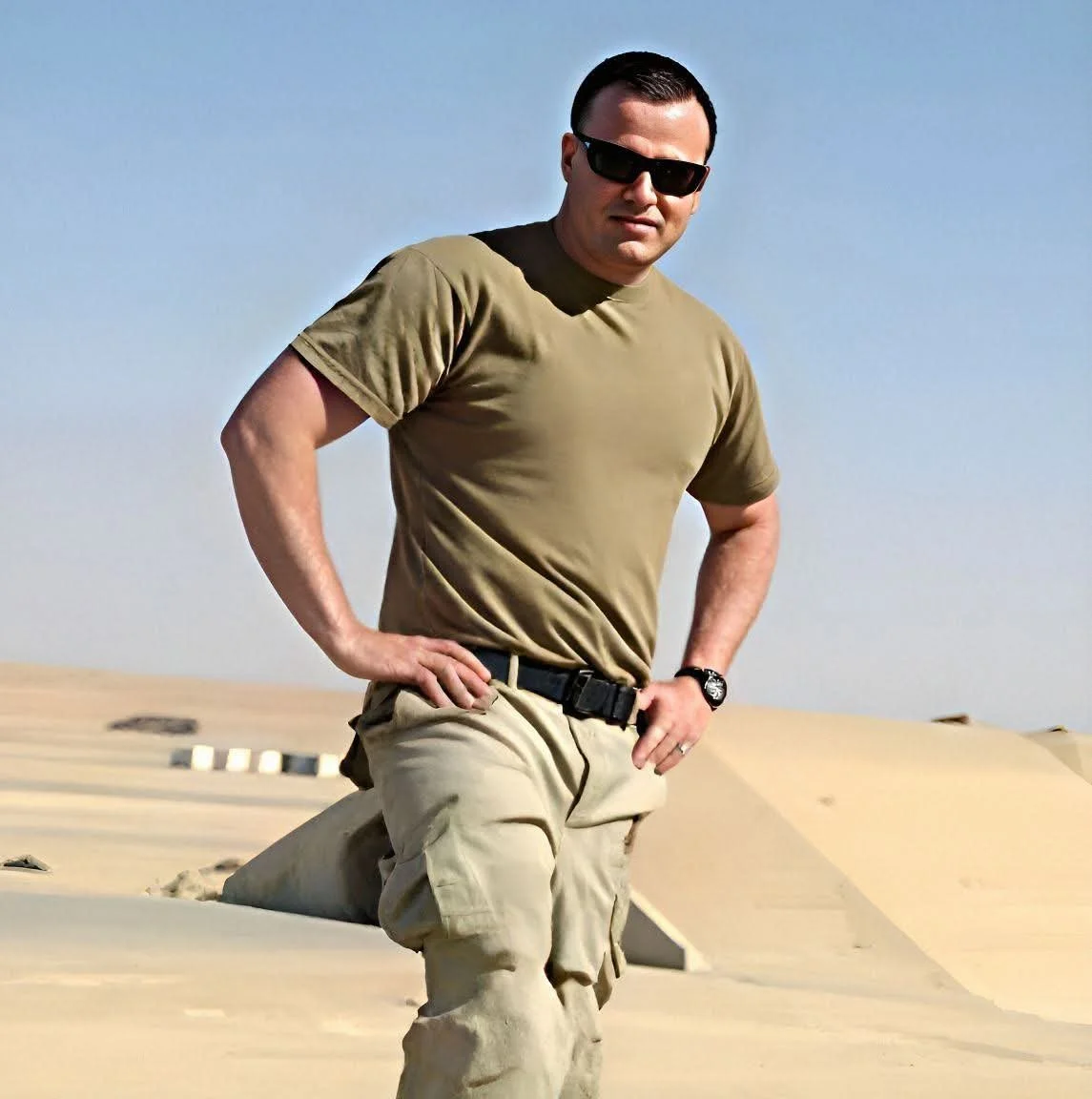 A man wearing sunglasses, a khaki T-shirt, and cargo pants standing outdoors on a hill with a clear sky.