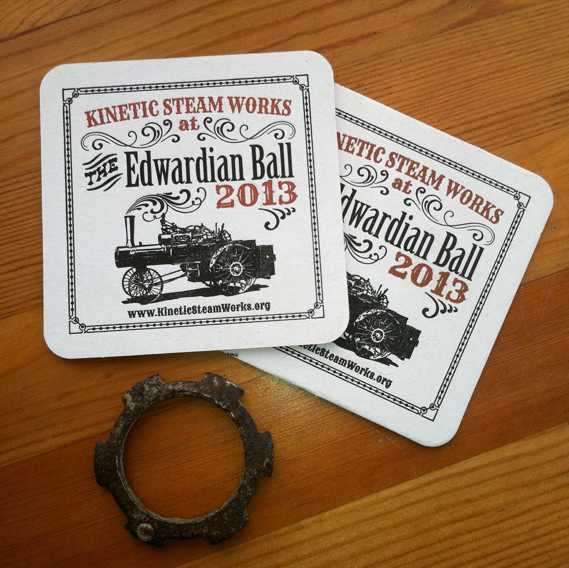 Custom designed two color letterpress coasters with a vintage steam tractor graphic and decorative border on a wooden surface, with a rusty gear nearby.