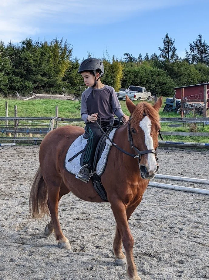 A young boy wearing a helmet riding a brown horse with a white stripe on its face in an outdoor riding arena.