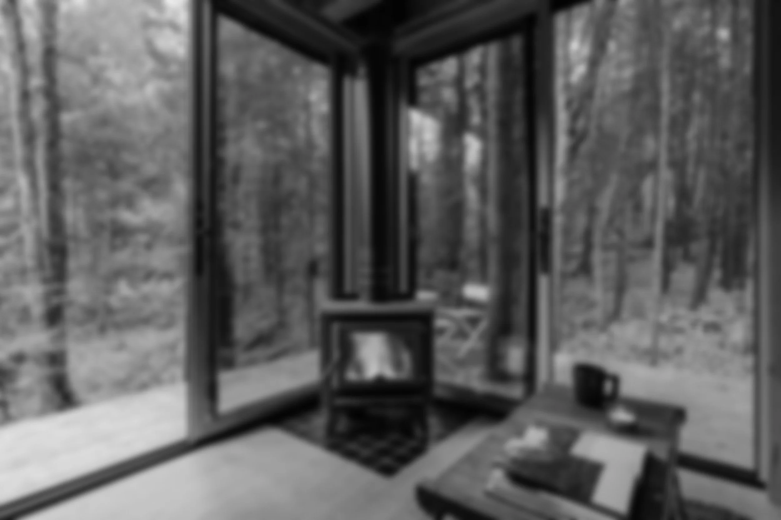 Black and white photo of a cozy room with large windows showing a forest outside, a small wood stove in the corner, and a wooden table with a cup and some papers.