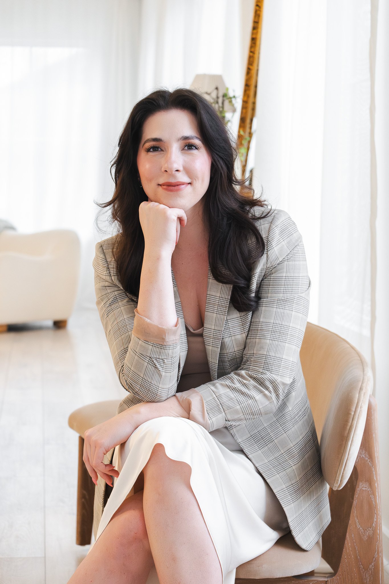 A woman with dark hair, wearing a beige checked blazer and light-colored skirt, sitting on a chair with her chin resting on her hand, in a well-lit room with white curtains and a measuring tape in the background.