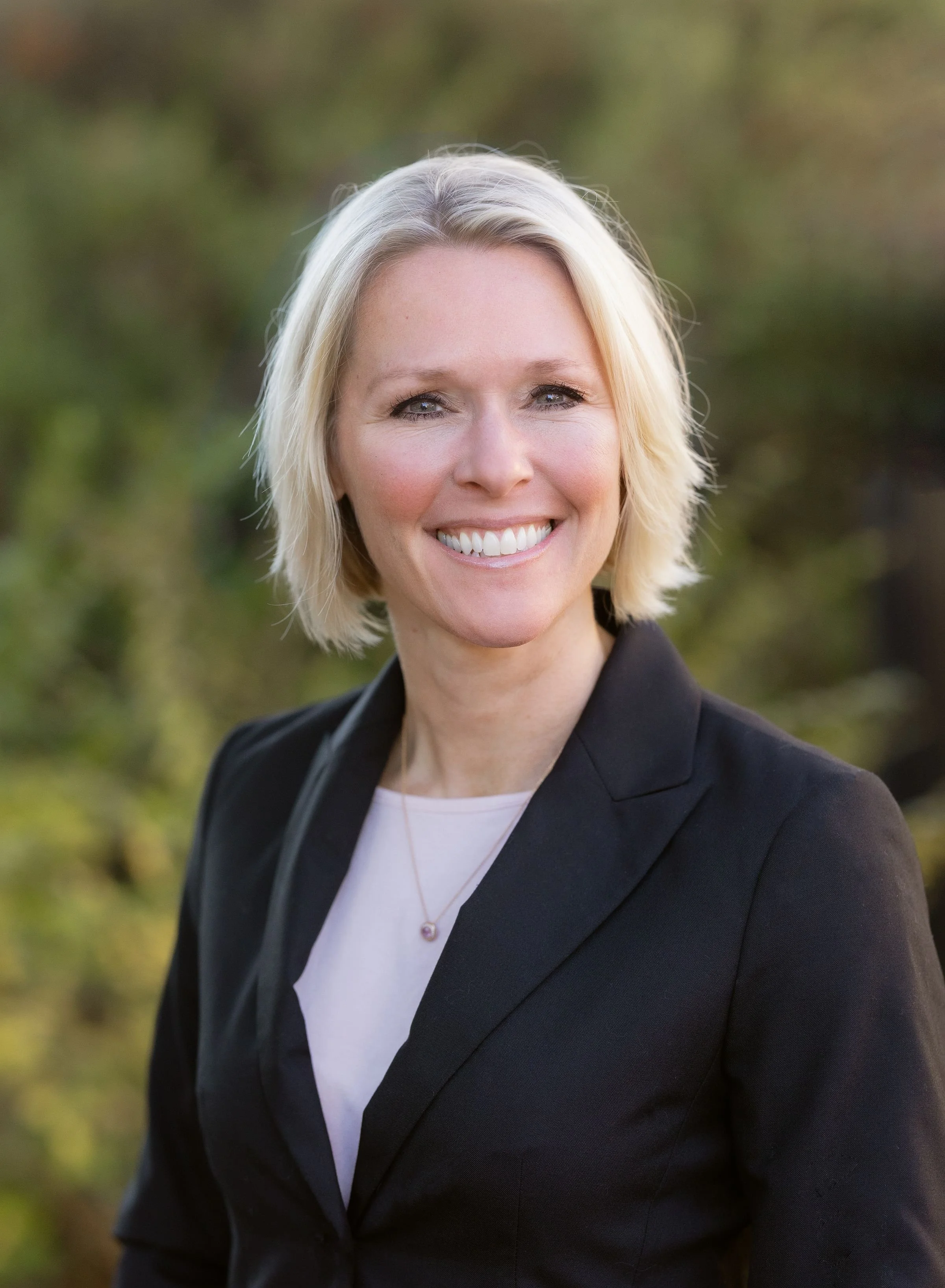 A woman with short blonde hair smiling outdoors with a blurred green background.