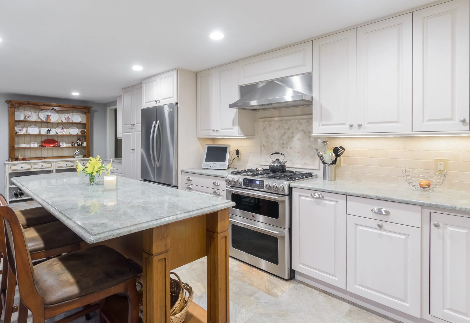 Kitchen with white cabinets, stainless steel refrigerator, stove, microwave, and a kitchen island with a gray countertop. There's a wooden hutch with plates and decorative items in the background, Rooms With A View Tile