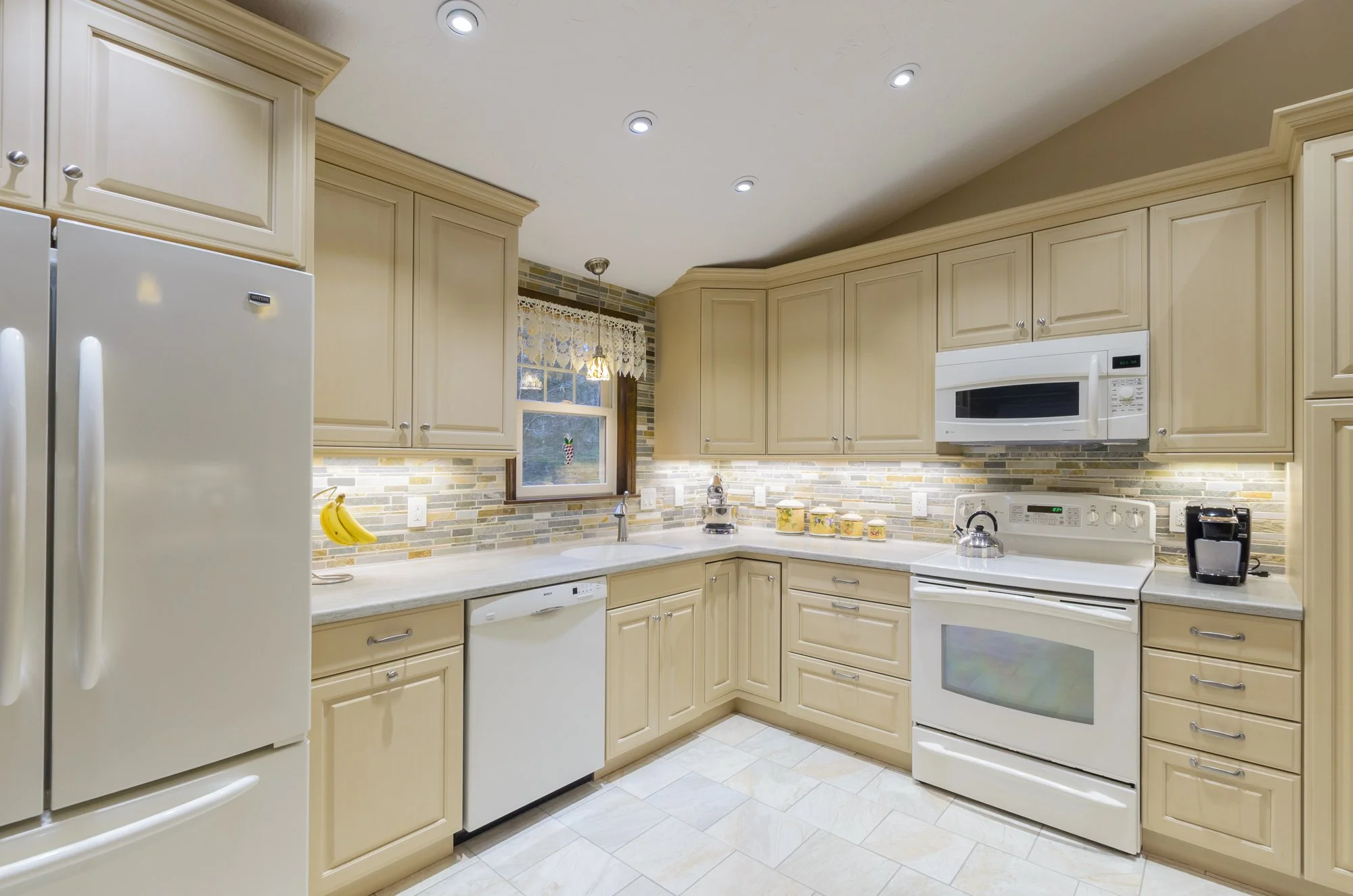 Kitchen with cream-colored cabinets, white appliances, granite countertop, tile backsplash, window with lace curtain, and recessed ceiling lights. Rooms With A View Tile