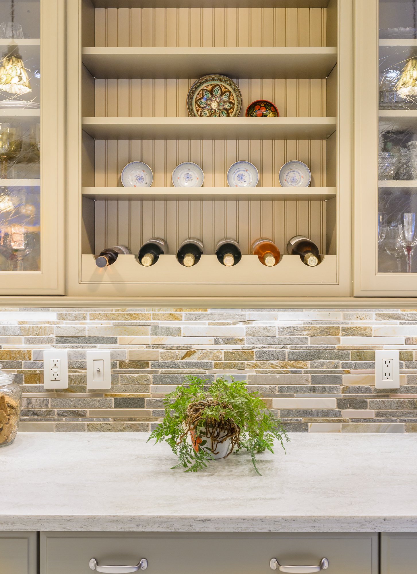 Kitchen cabinet with glasses and plates, wine bottles resting in a rack, decorative plates on the shelf, potted plant on the counter, and a brick backsplash. Rooms With A View Tile