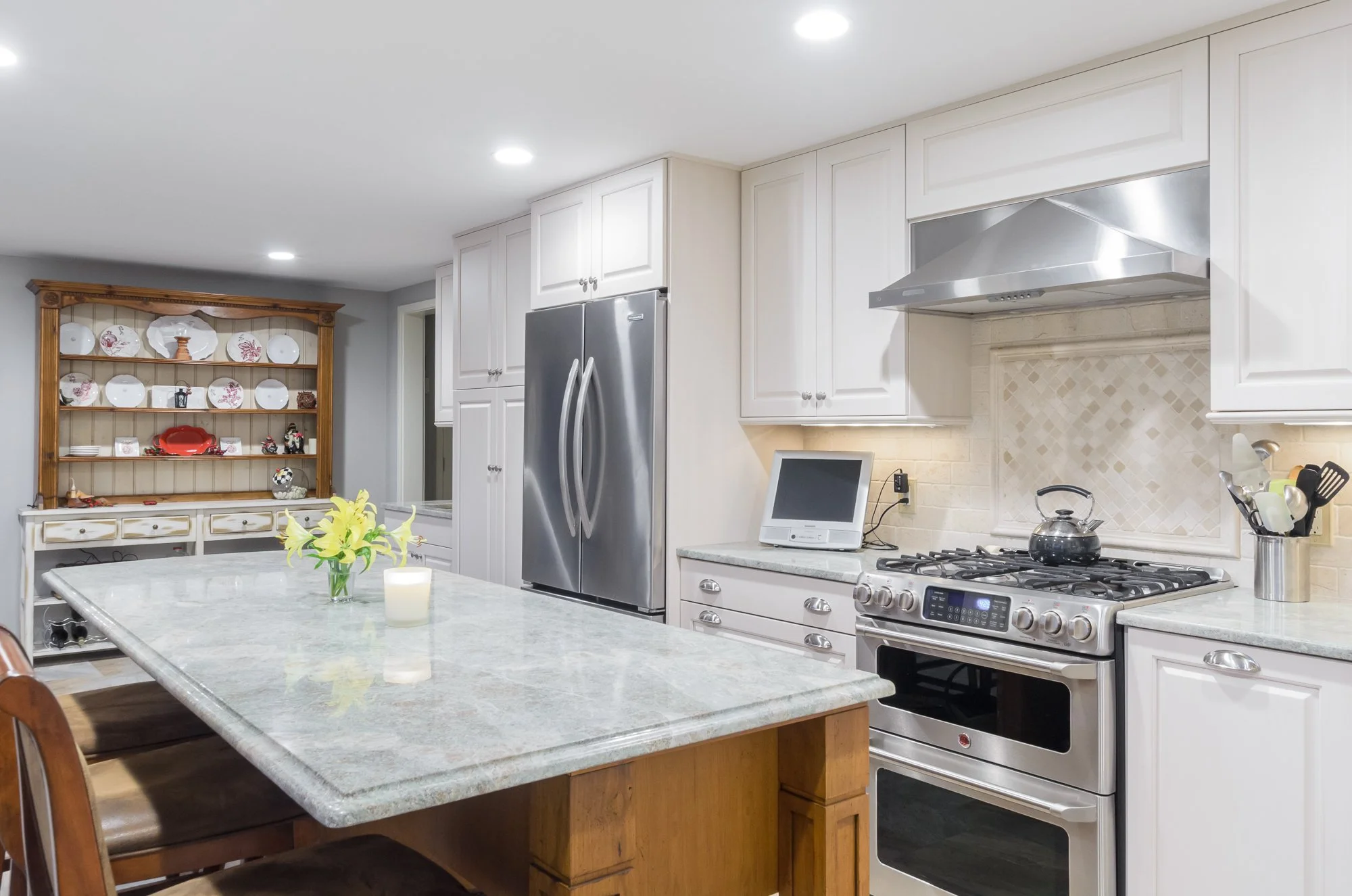 Modern kitchen with white cabinets, stainless steel refrigerator and stove, marble countertop island, and a shelf with decorative plates in the background. Rooms With A View Tile