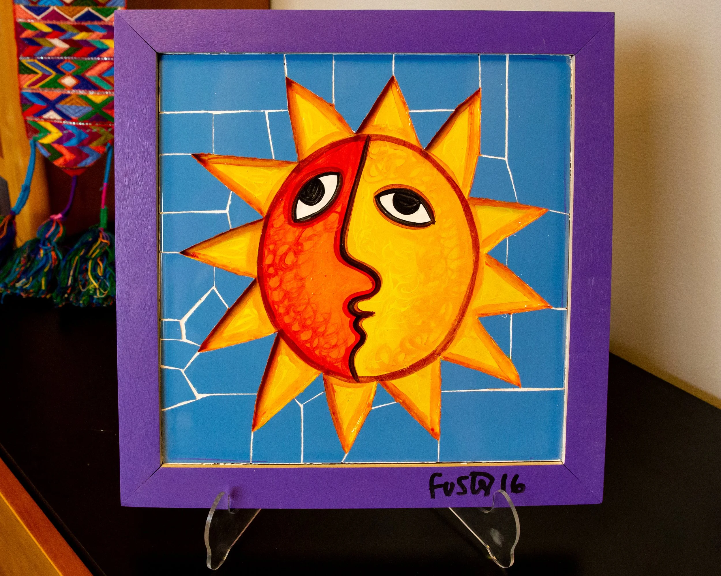A colorful mosaic artwork depicts a sun with a face, featuring a yellow and orange face with expressive eyes and a black outline, surrounded by bright orange sun rays on a blue background, framed in purple.