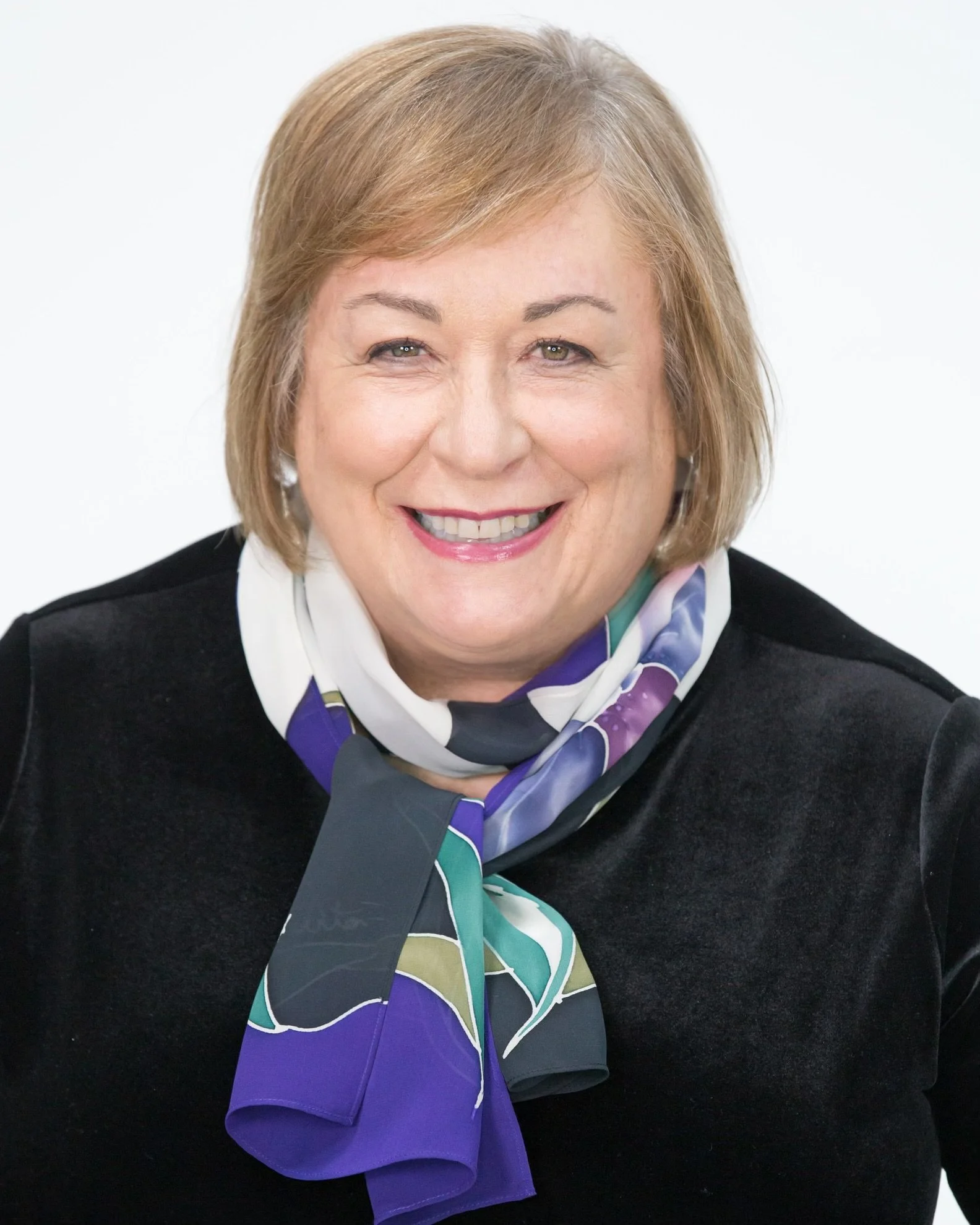 Naomi Perman Attorney at Law, smiling woman with light brown hair, wearing a black top and a colorful scarf, posing against a white background.