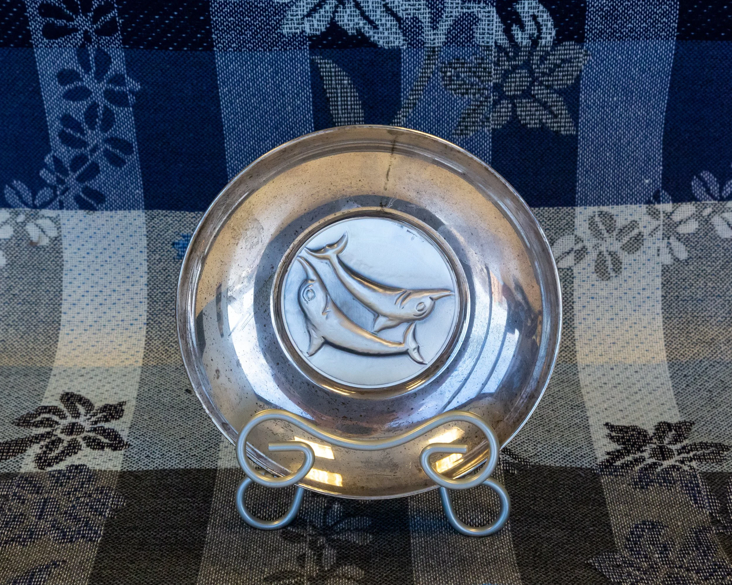 Round silver serving tray with embossed dolphin design, placed on a checkered tablecloth with floral patterns, and held by a metal stand.