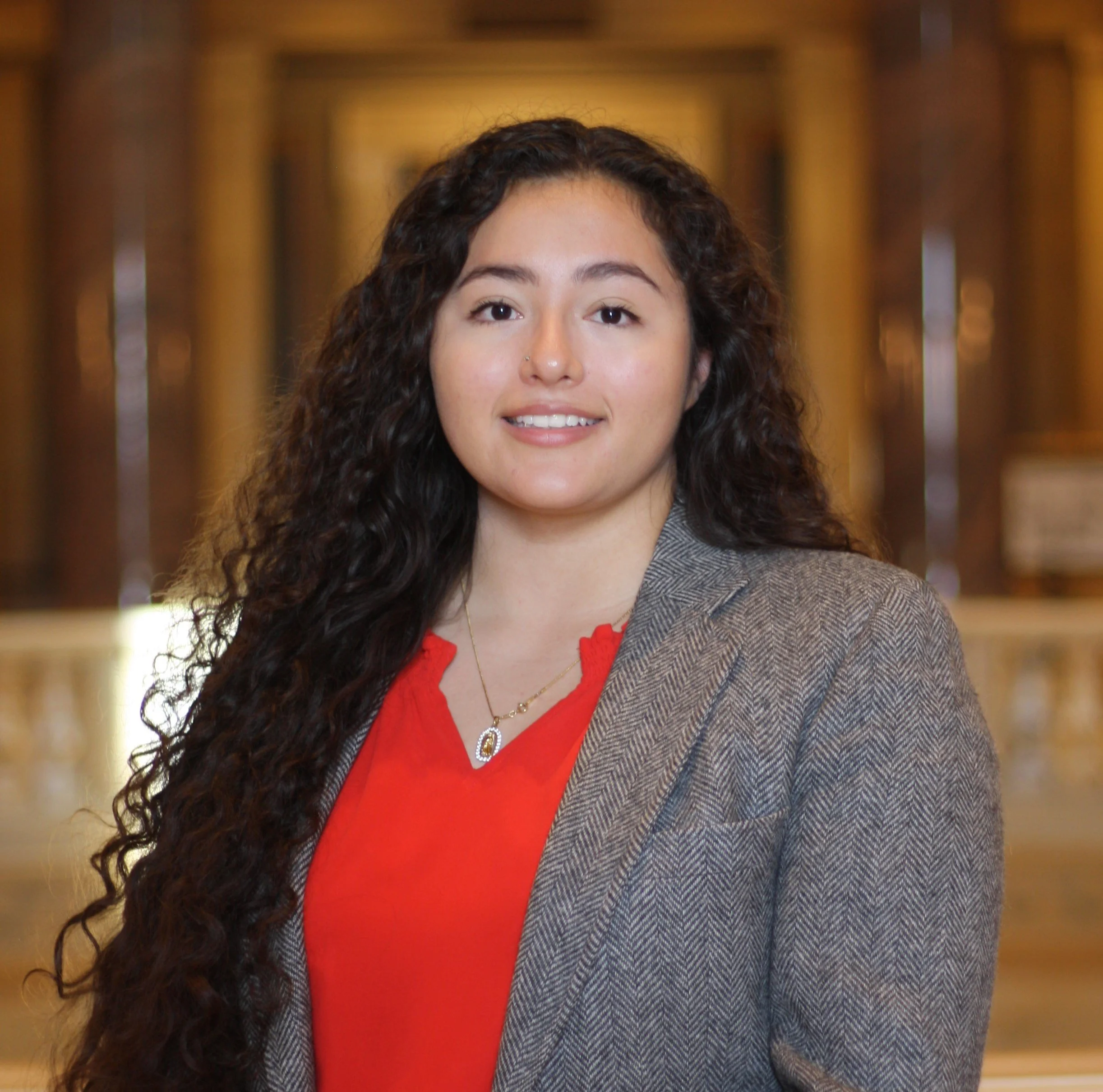 Alejandra Kelly  Legal Assistant, in Minnesota,A woman with long curly dark hair, wearing a gray blazer over a red blouse, standing in a wooden-paneled room.