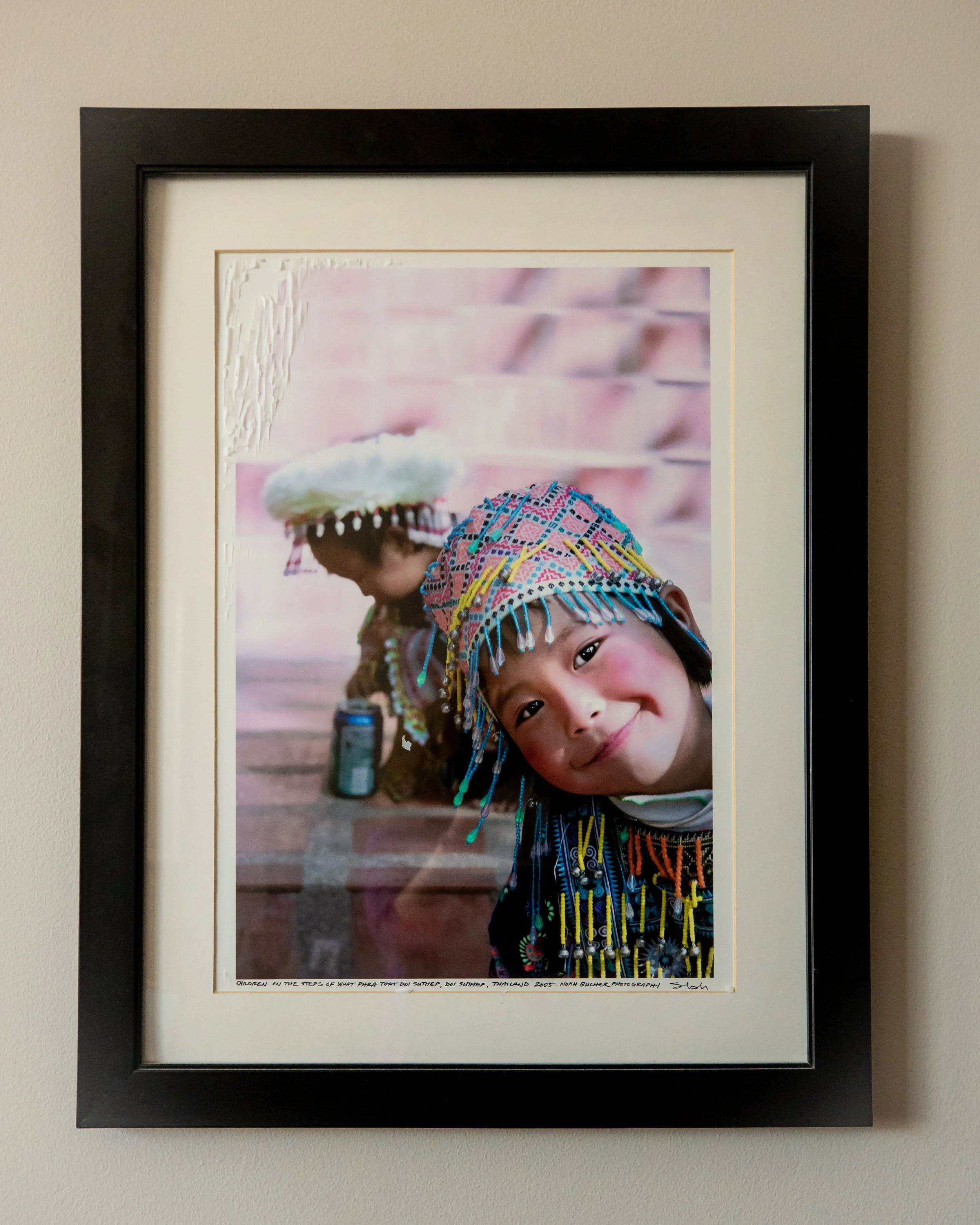 Photograph of two children in traditional ethnic attire with colorful beaded accessories, one smiling at the camera, the other in the background, against a pinkish backdrop.