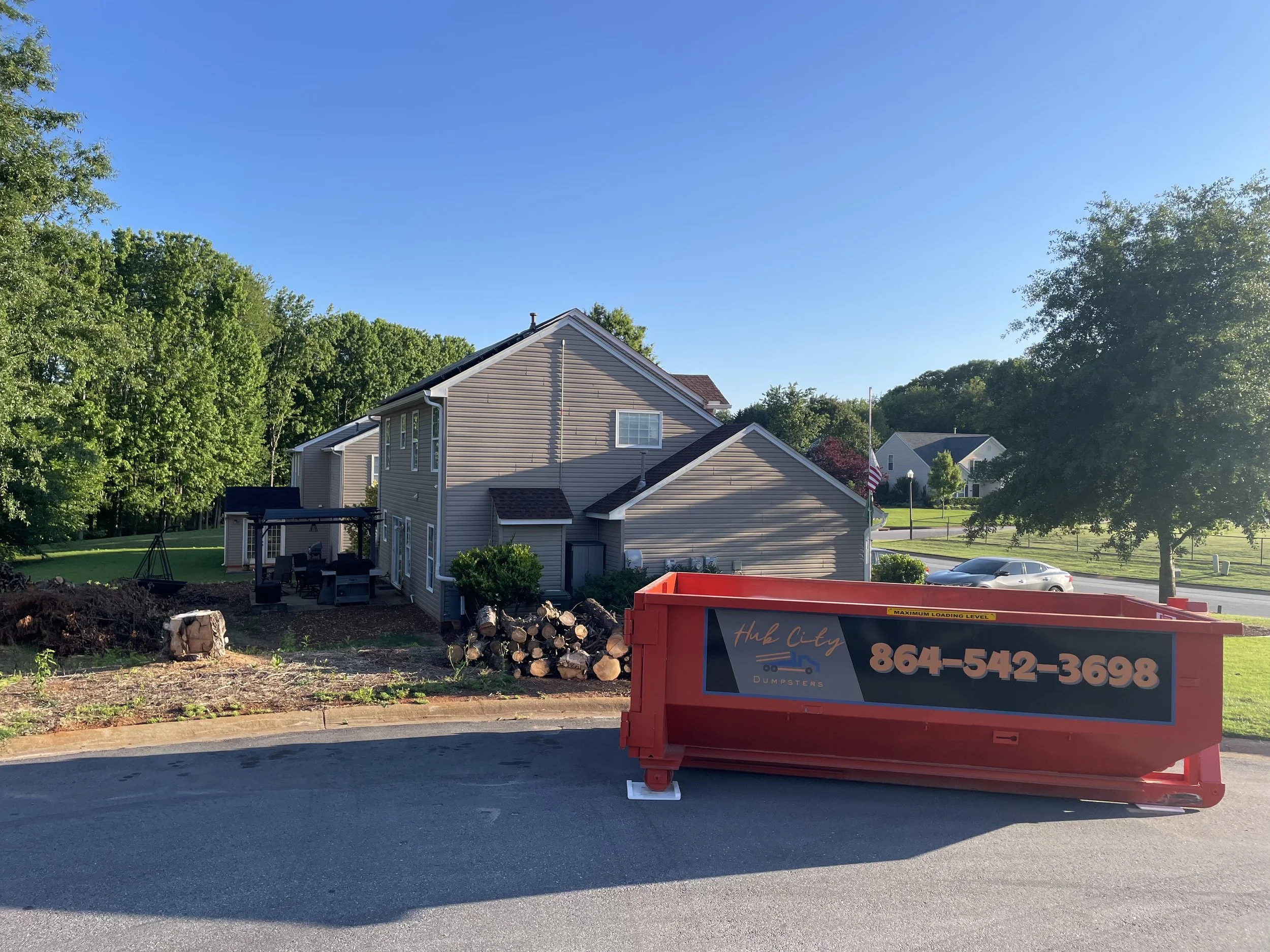 Residential house with a backyard, large trees, a pile of firewood, a swing set, and a red dumpster with the text "Hub City Dumpsters" and a phone number in front of the house.