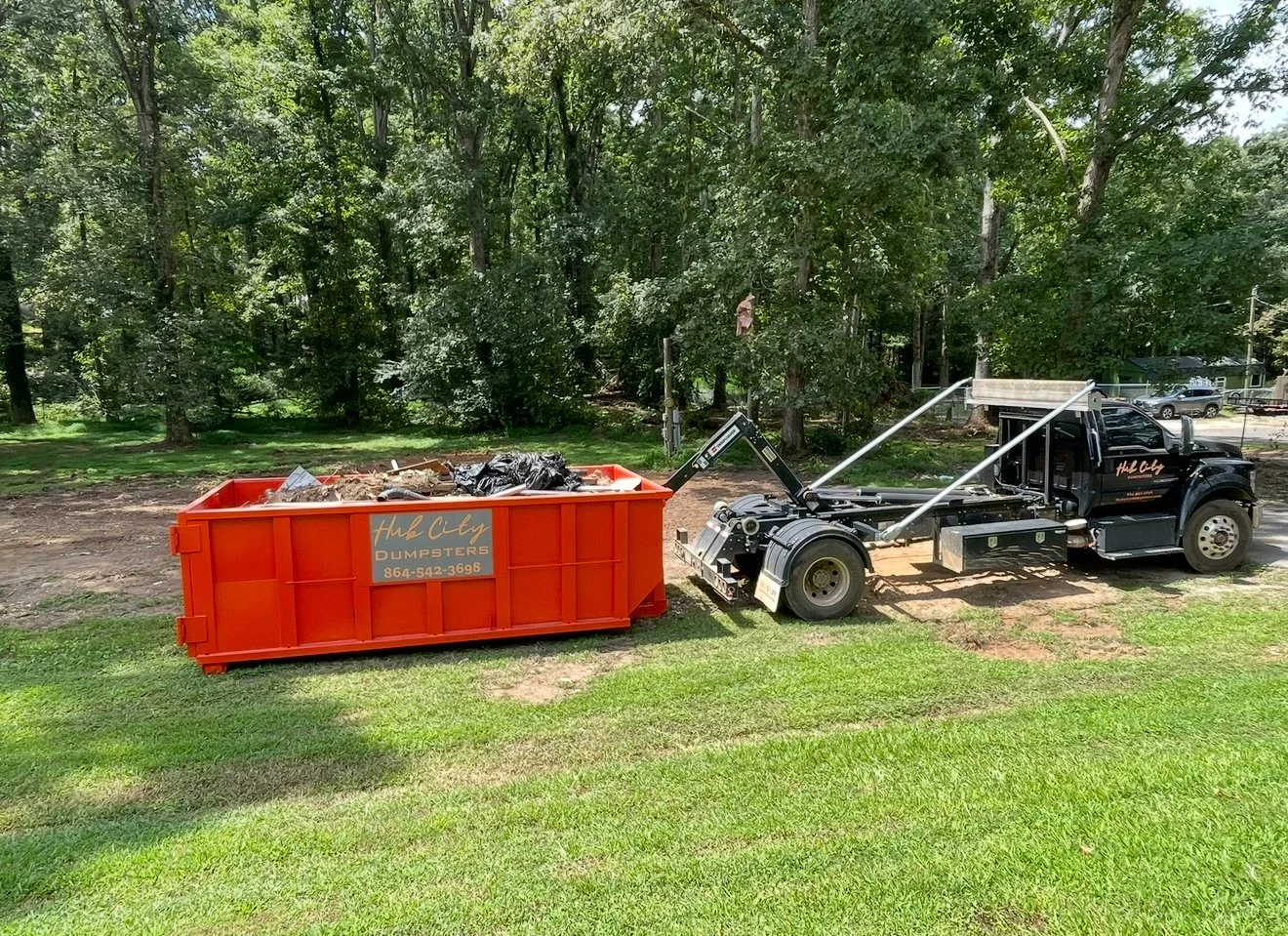A large orange dumpster filled with trash and debris, with a black truck nearby, in a green outdoor area with trees in the background.
