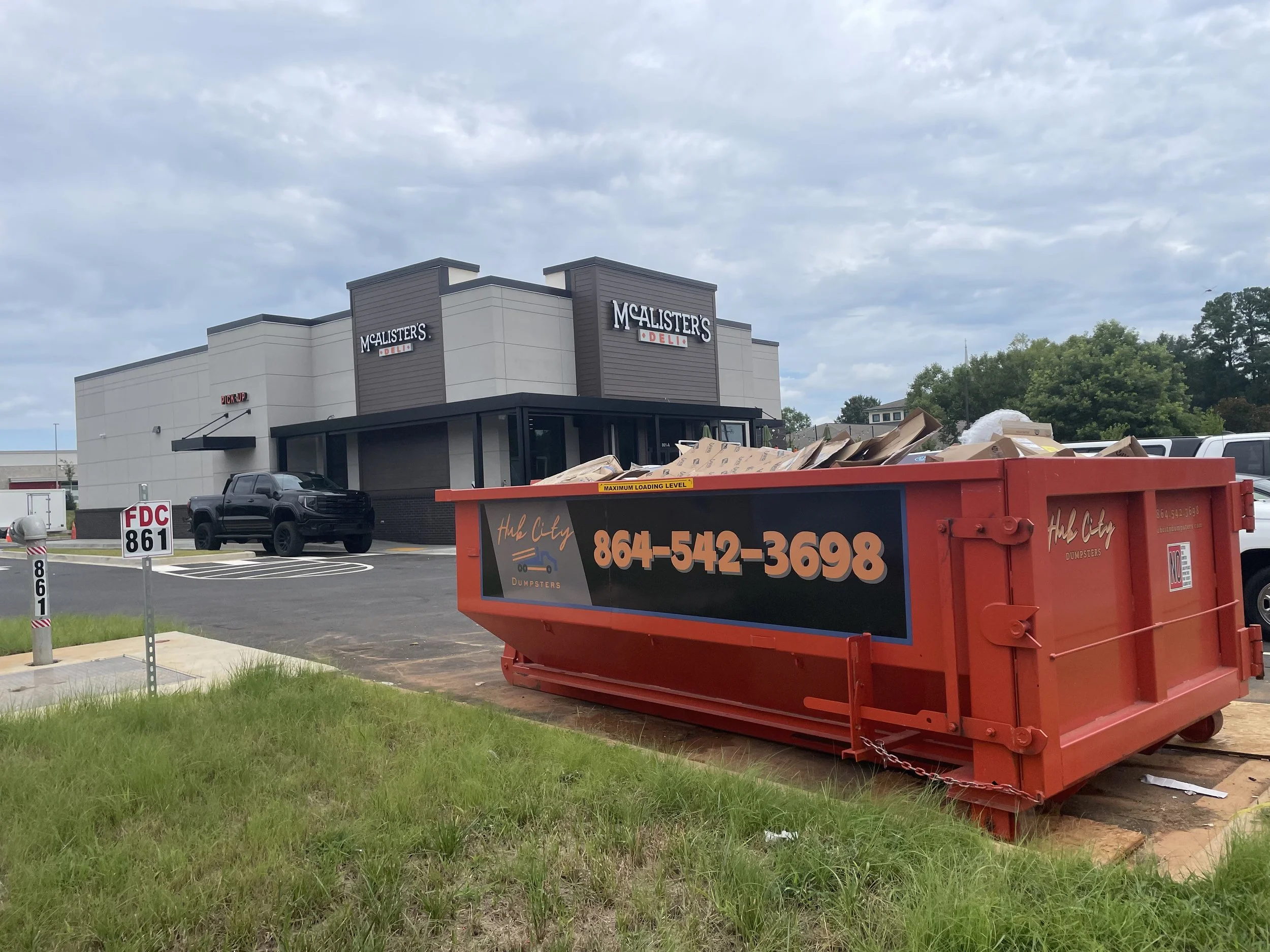A McAlister's Deli restaurant building with a dumpster in the foreground filled with cardboard and waste, a parked black pickup truck, and a parking lot under cloudy skies.