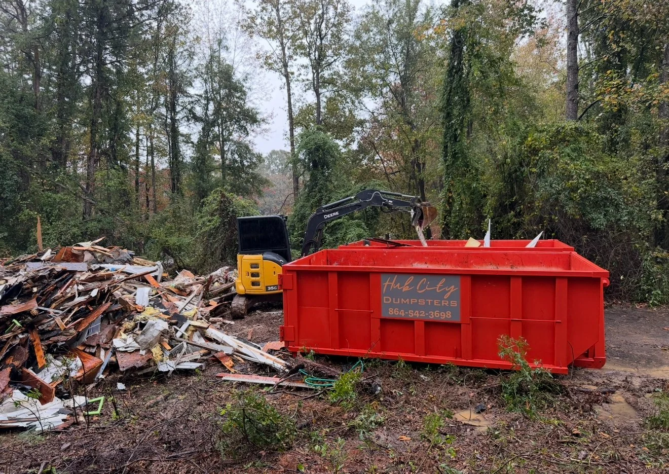 A small yellow and black excavator with a black arm next to a large orange dumpster with the sign 'Hub City Dumpster' and a phone number, in a wooded outdoor area with debris and piles of scrap wood and metal.