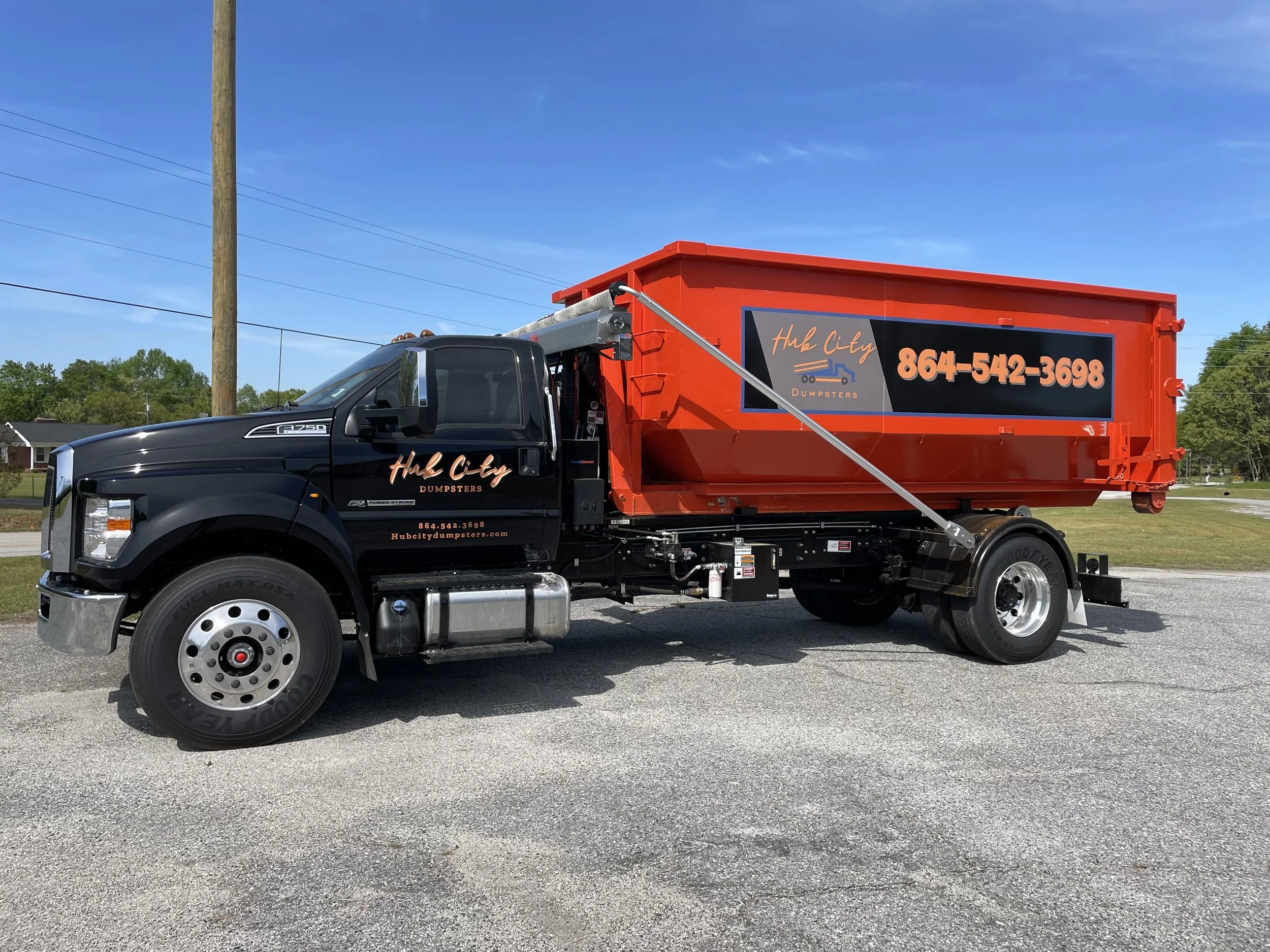 A black and red dumpster truck parked on a gravel surface under a blue sky, with trees and a utility pole in the background. The truck has the company name 'Hub City Dumpsters' and phone number '864-542-3698' displayed on the side.