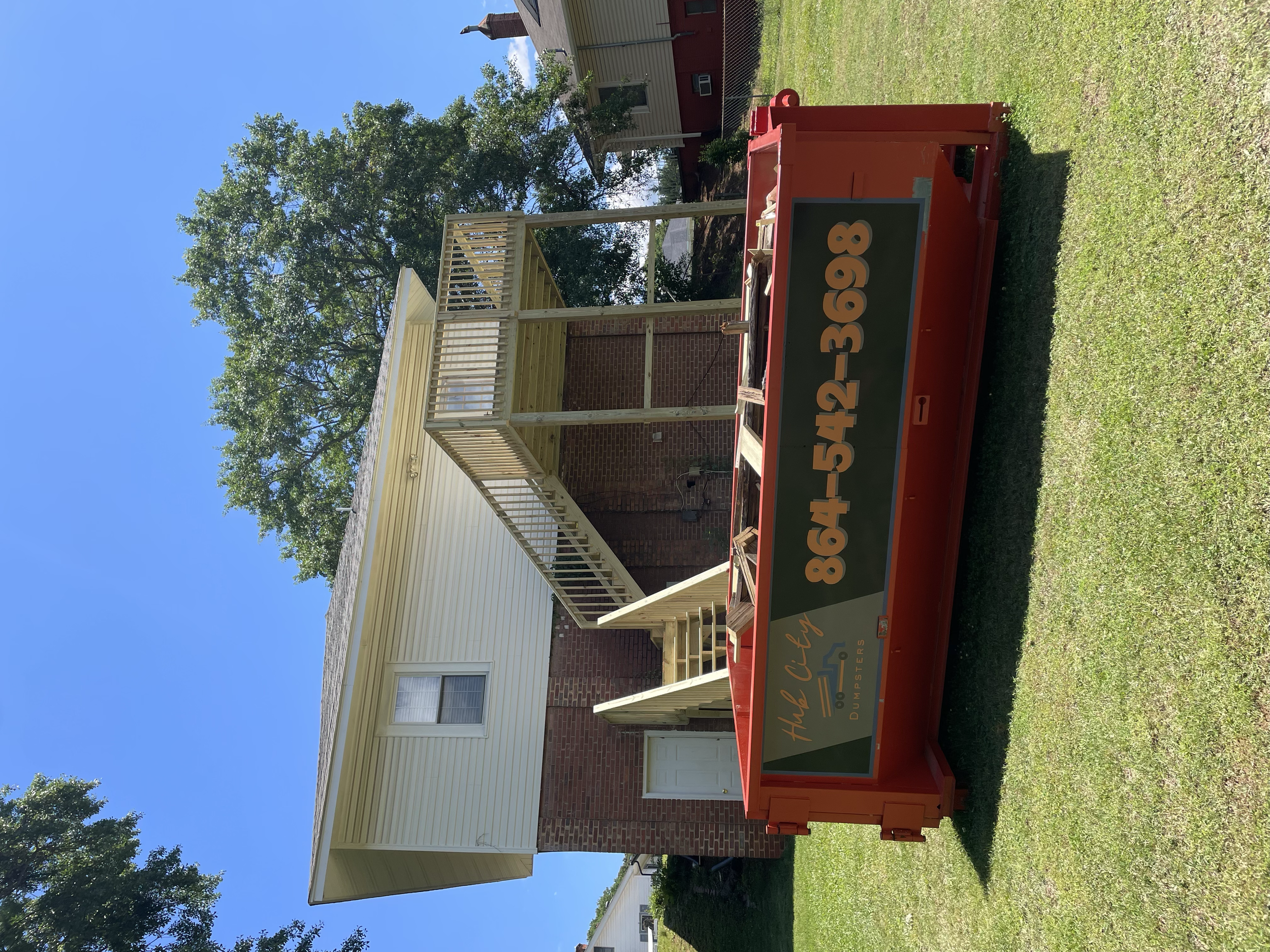 A red dumpster with the sign 'Haul Dude Dumpsters' and phone number 864-542-3668 parked in front of a house with a yellow upper floor and brick lower wall, and a small porch with stairs. The house is surrounded by green grass and trees under a clear 