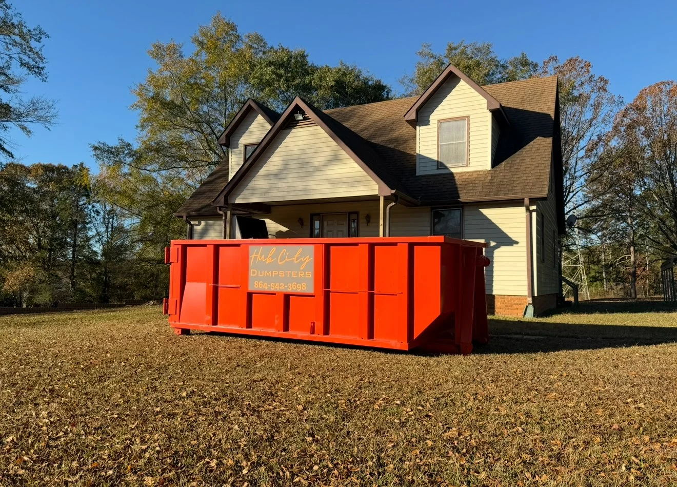 Red dumpster in front of a two-story house with beige siding and multiple gabled roofs, surrounded by trees with fall foliage.