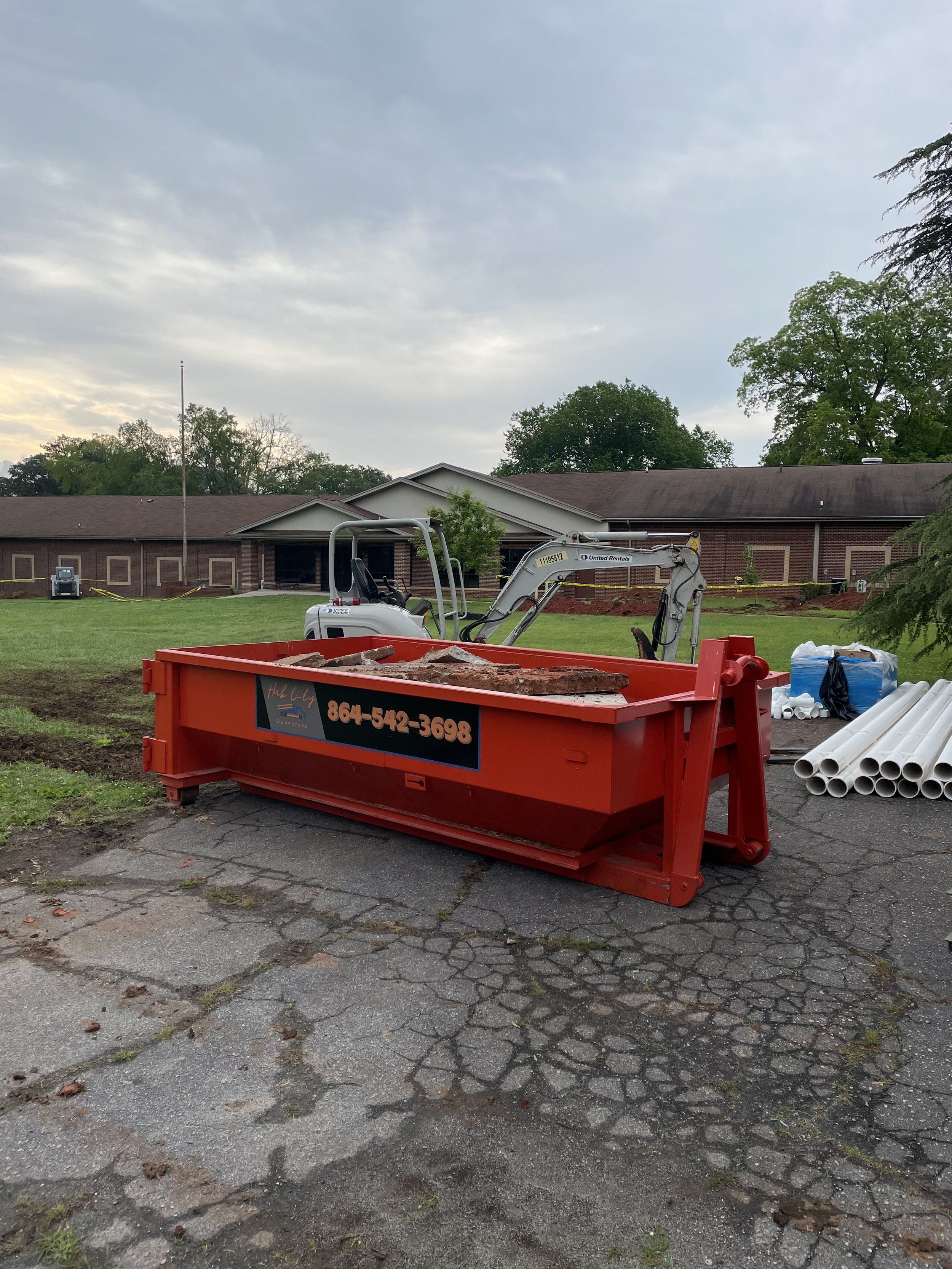 Construction equipment including a large orange dumpster filled with dirt, a small white mini excavator, and various pipes and materials in front of a building with a lawn and cloudy sky.
