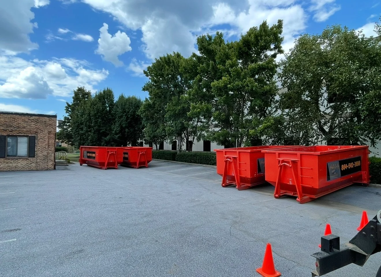 Empty parking lot with large red dumpsters, small orange traffic cones, and a brick building with trees and a blue sky with clouds in the background.