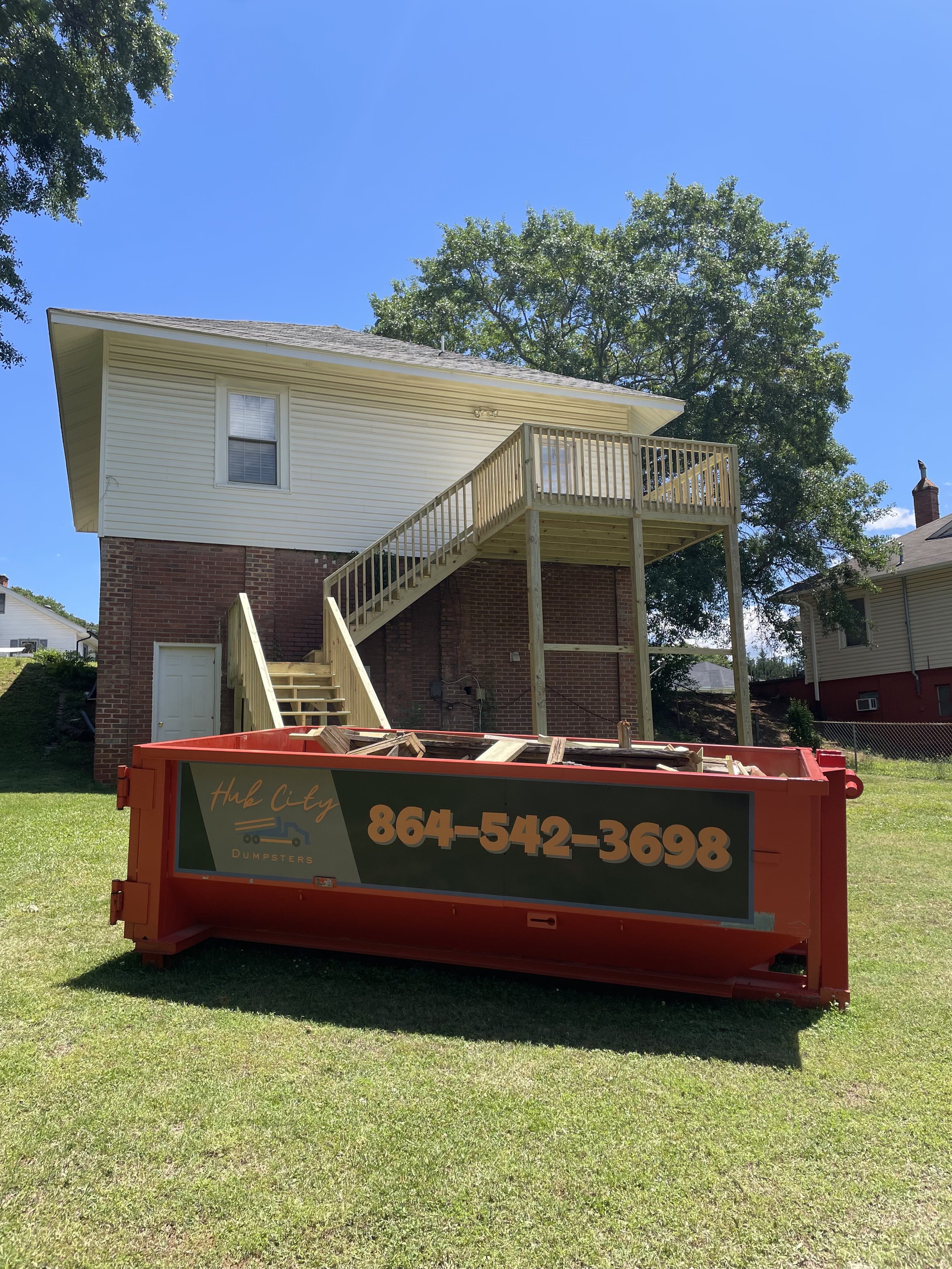 New wooden deck being built on a brick house with stairs leading up to it. A red dumpster with the phone number 864-542-3698 and sign 'Hub City Dumpster' is in the foreground. There are trees and neighboring houses in the background on a sunny day.