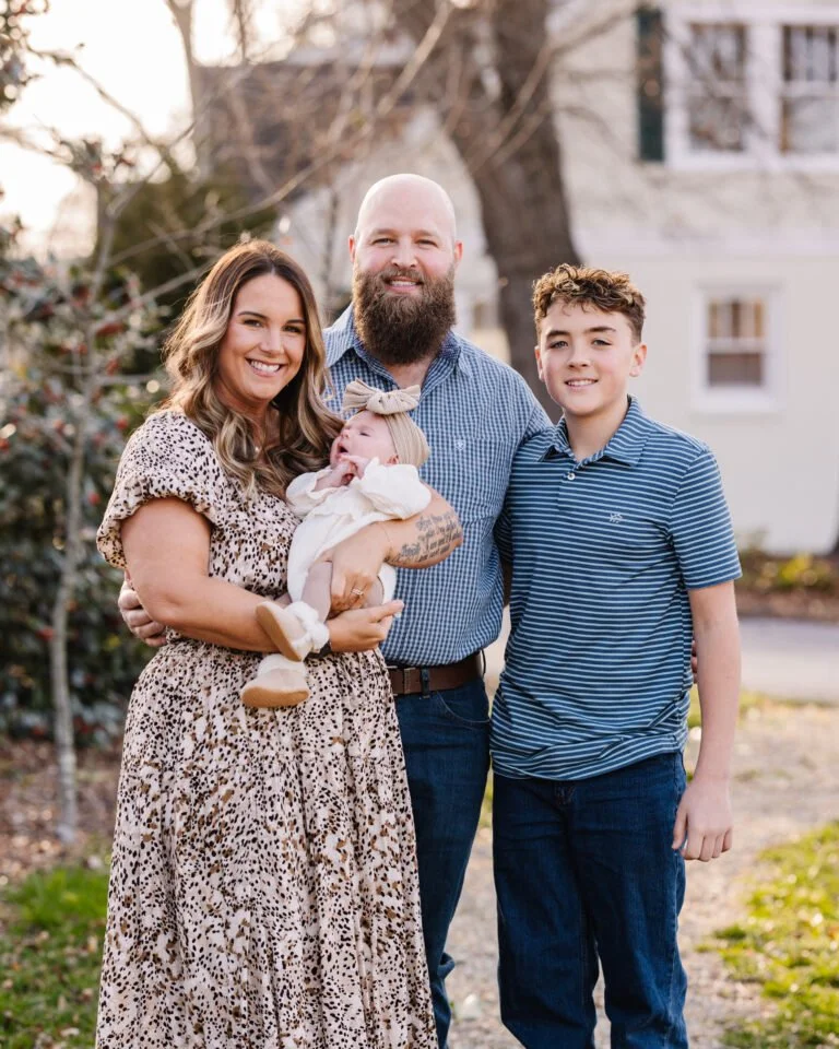 A family of four outdoors, smiling, standing close together on a lawn with a house and trees in the background. The woman is holding a small baby girl.