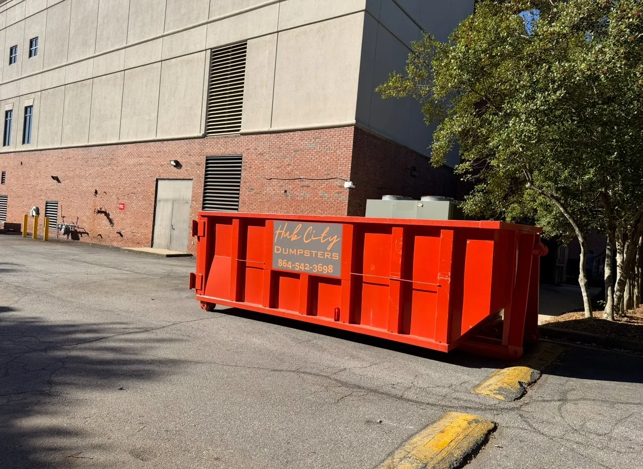 A large red dumpster with a sign reading 'Hub City Dumpsters' and a phone number, placed on a paved parking lot near a brick and concrete building with trees in the background.