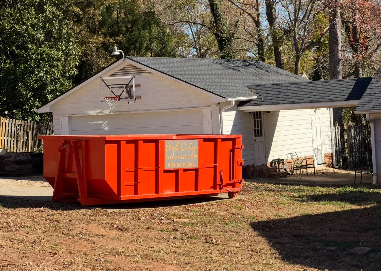 A residential garage with a basketball hoop above the garage door and an orange dumpster in the front yard near the house. The house has white siding and a gray roof, with trees in the background.