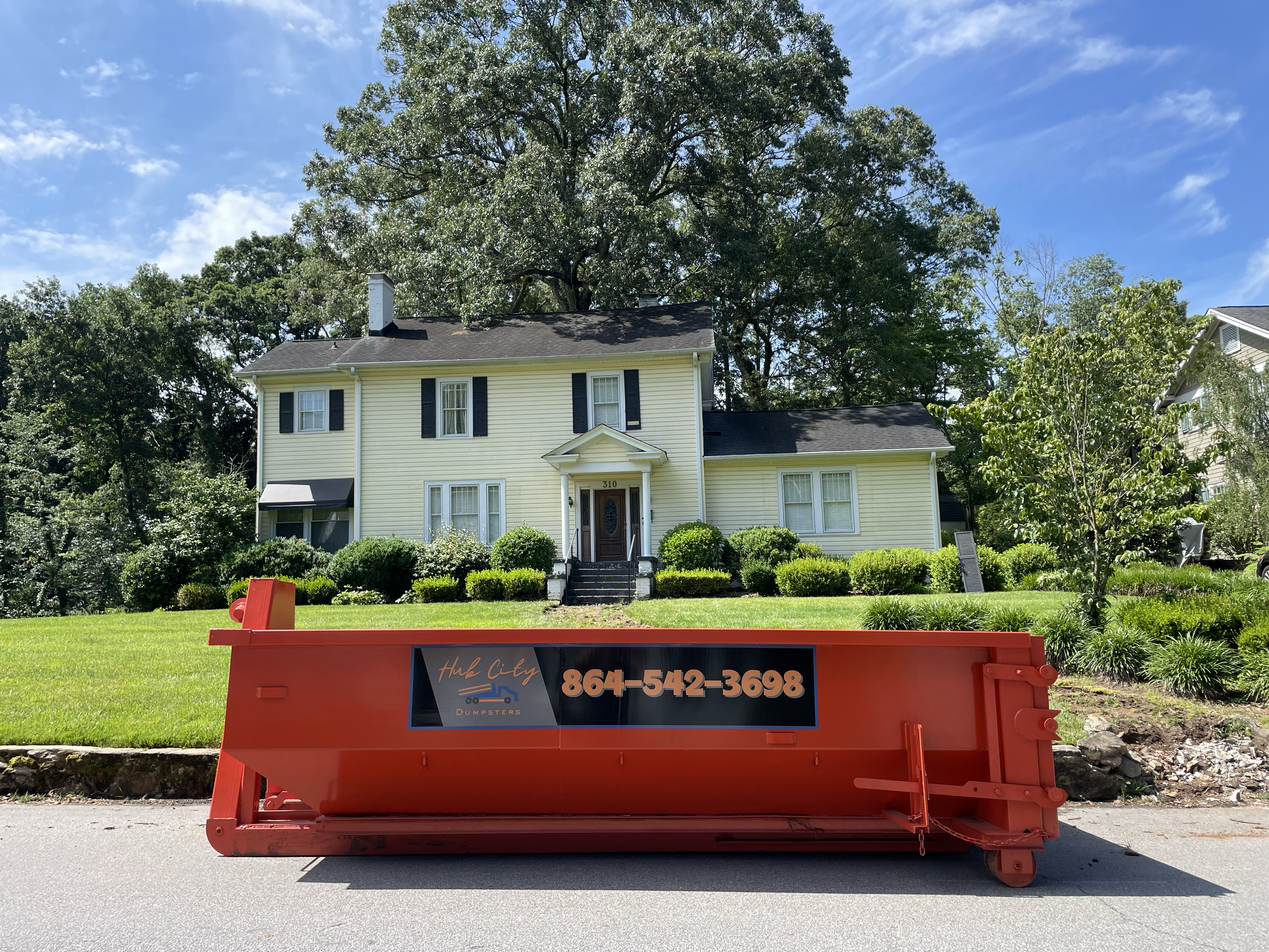 A residential house with a yellow exterior and black shutters, surrounded by greenery, with a large tree behind it. In the foreground, a bright red dumpster with the company name 'Hub City Dumpsters' and phone number 864-542-3698 is placed on the street.
