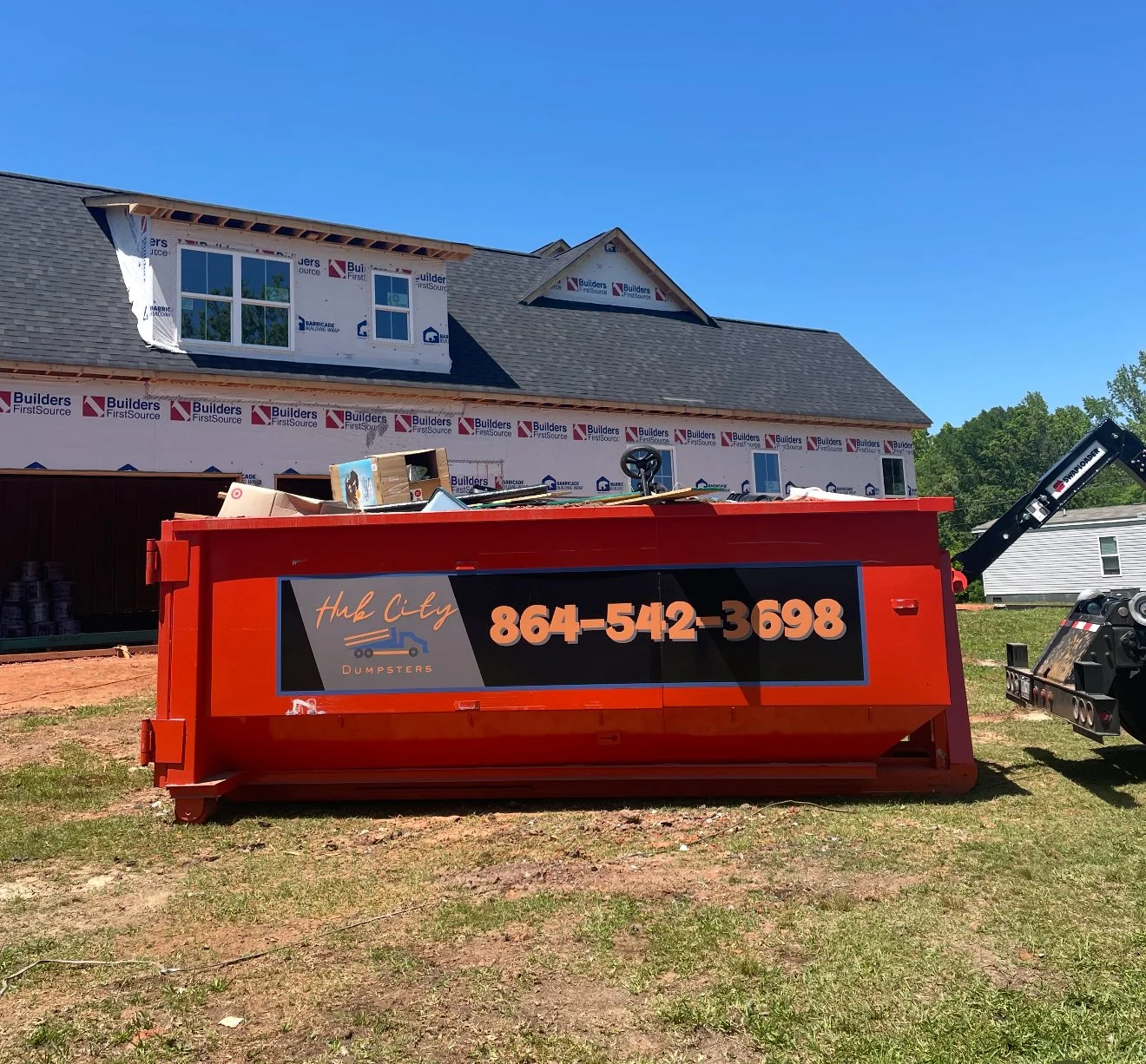 Red dumpster with Hub City Dumpsters contact information parked on a grassy area in front of a house under construction, with a blue sky above.