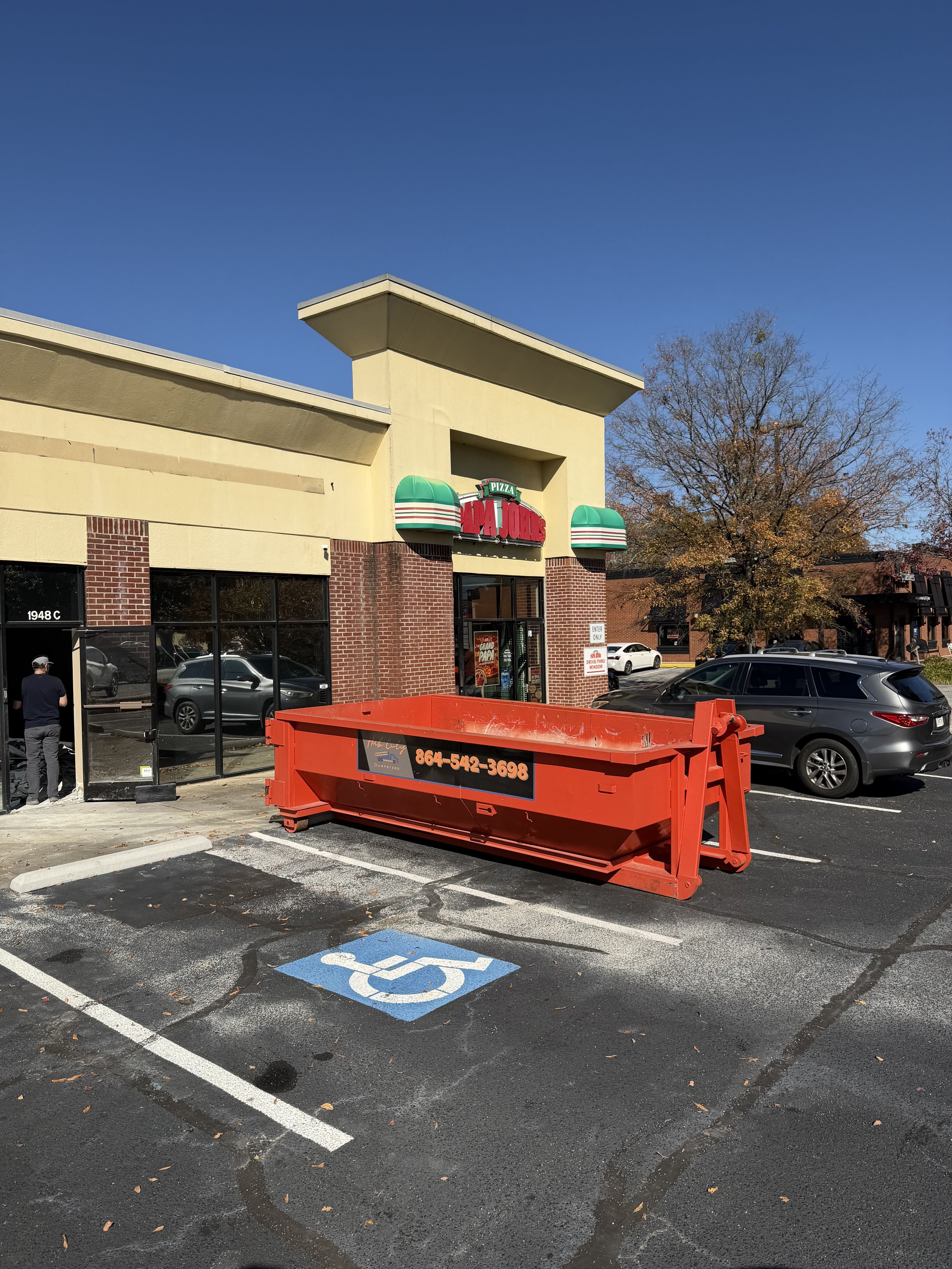 Parking lot in front of a Papa John's pizza restaurant with a large orange dumpster, cars parked, and a man standing near the windows on a sunny day.