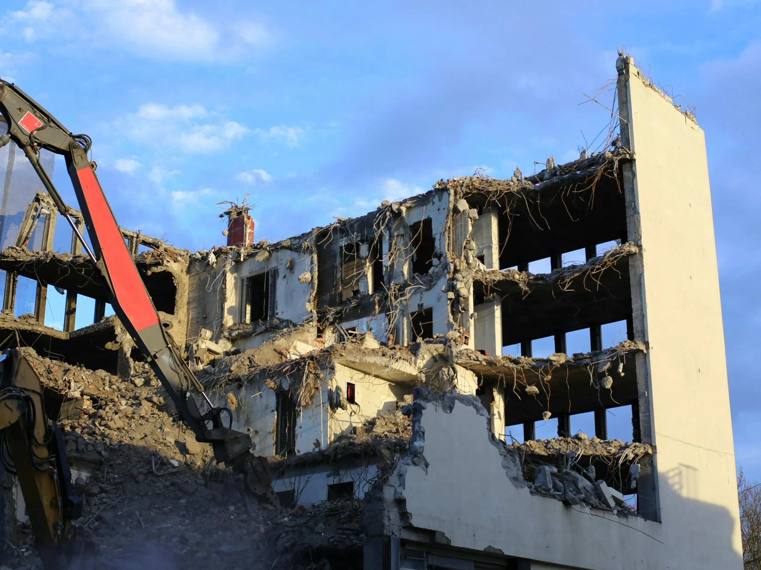 Partially demolished multi-story building with exposed interior and debris, including broken concrete and rebar, under a blue sky.