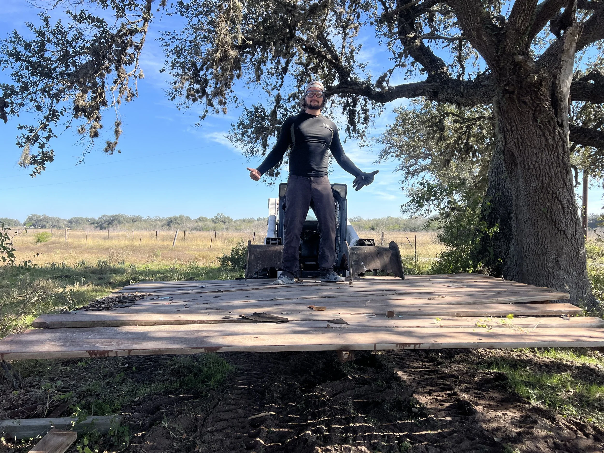 A man standing on a wooden bridge that is under construction, with a large tree and open field in the background under a clear blue sky.
