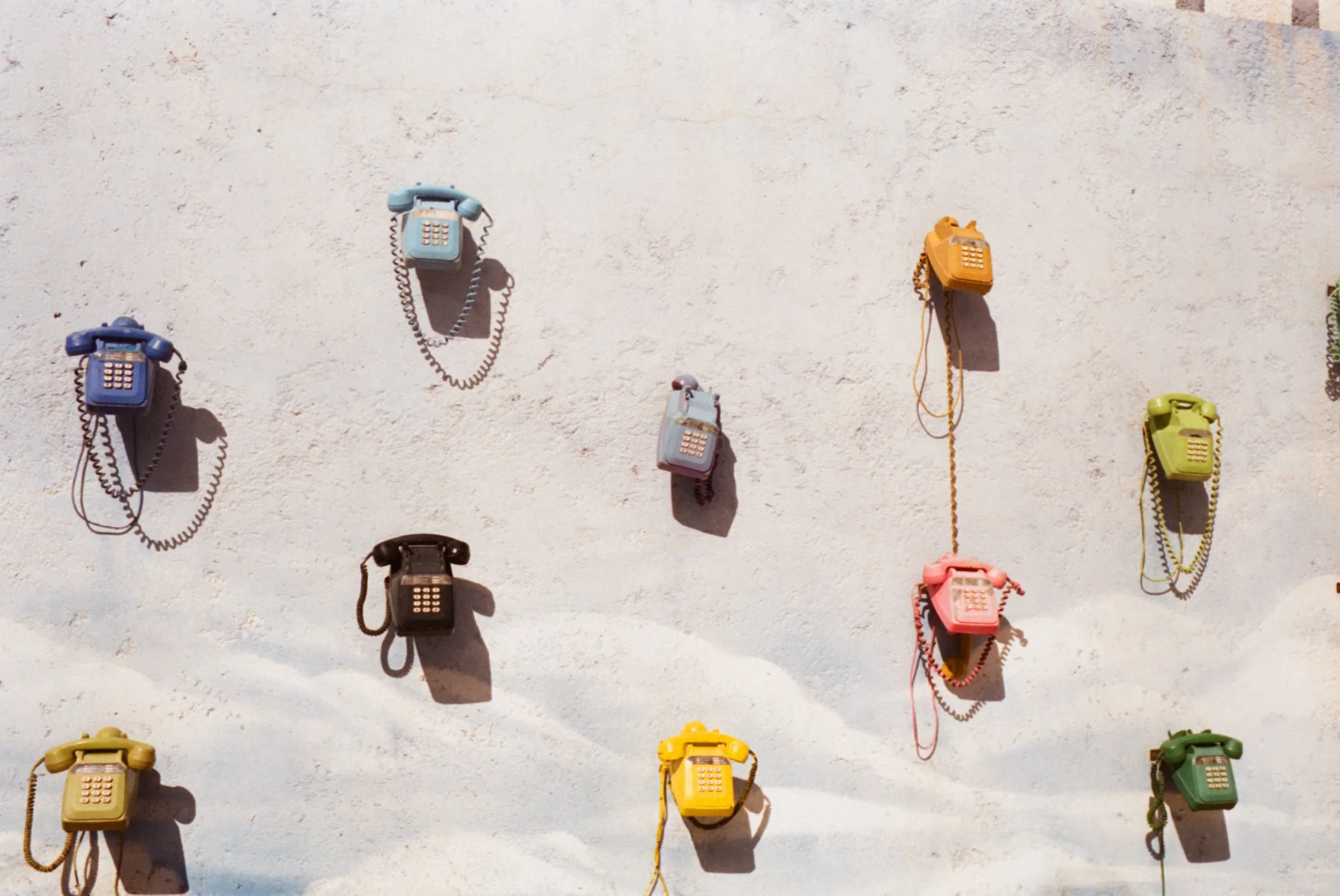 Multiple colorful vintage rotary phones mounted on a white wall, casting shadows.