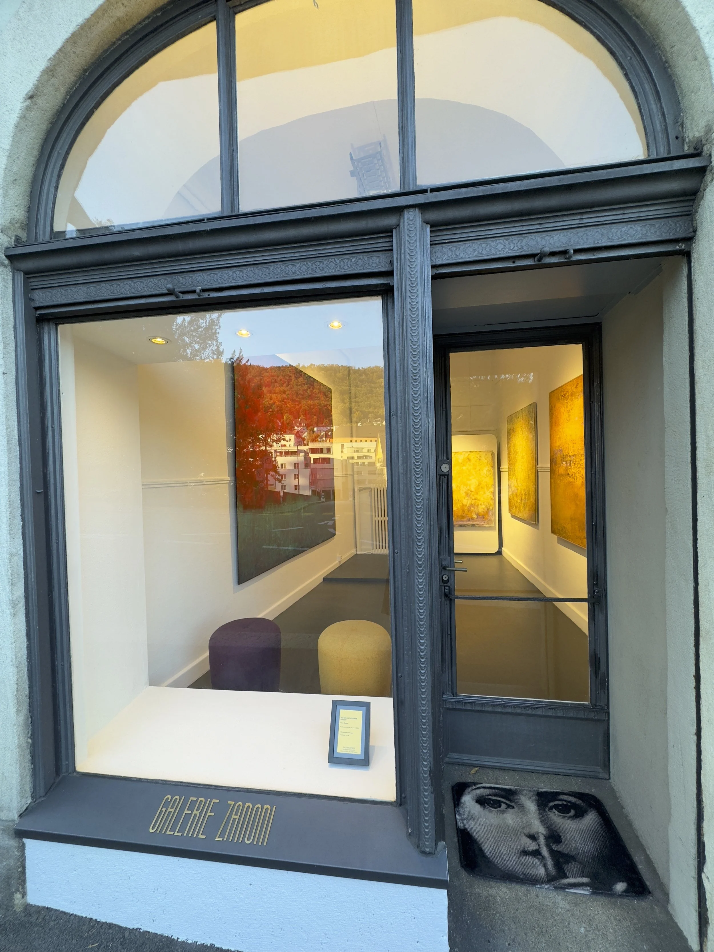 Auslage eines Galerieraums mit Kunstwerken an den Wänden, Sitzgelegenheiten und einer Begrüßungstafel, im Eingang eine schwarze Matte mit einem schwarzen und weißen Gesichtsfoto.