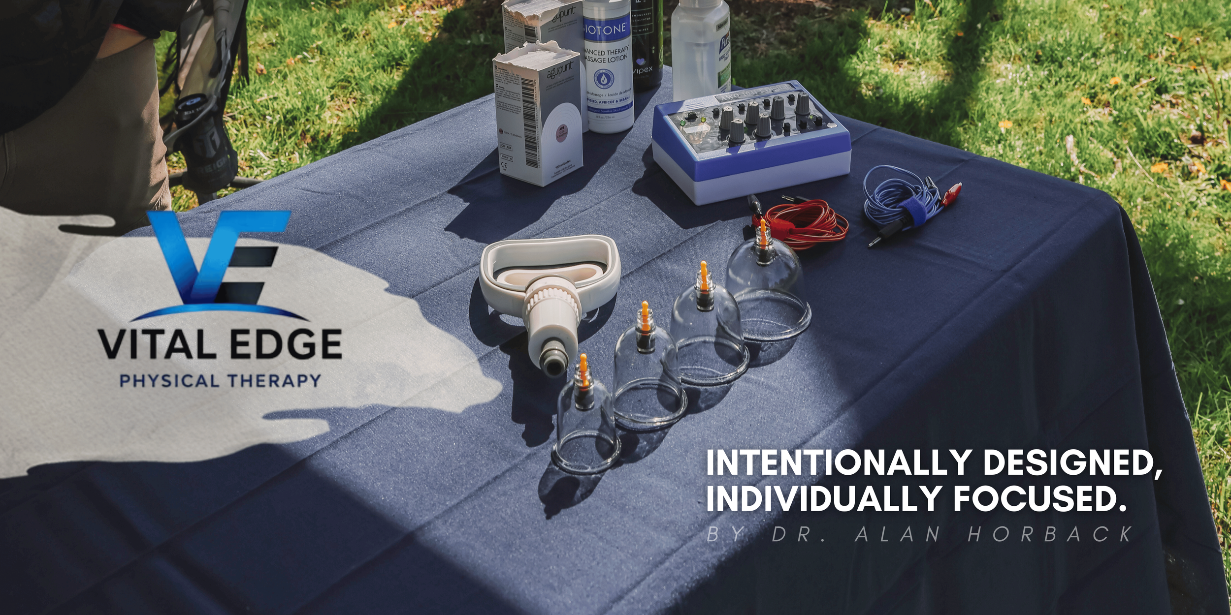 A table covered with a blue cloth outdoors, displaying various medical and therapy equipment including glass bottles, medical tape, a massage lotion, a small audio mixer, wires, and glass cups with thin tubes inserted, with grass and trees in the background.
