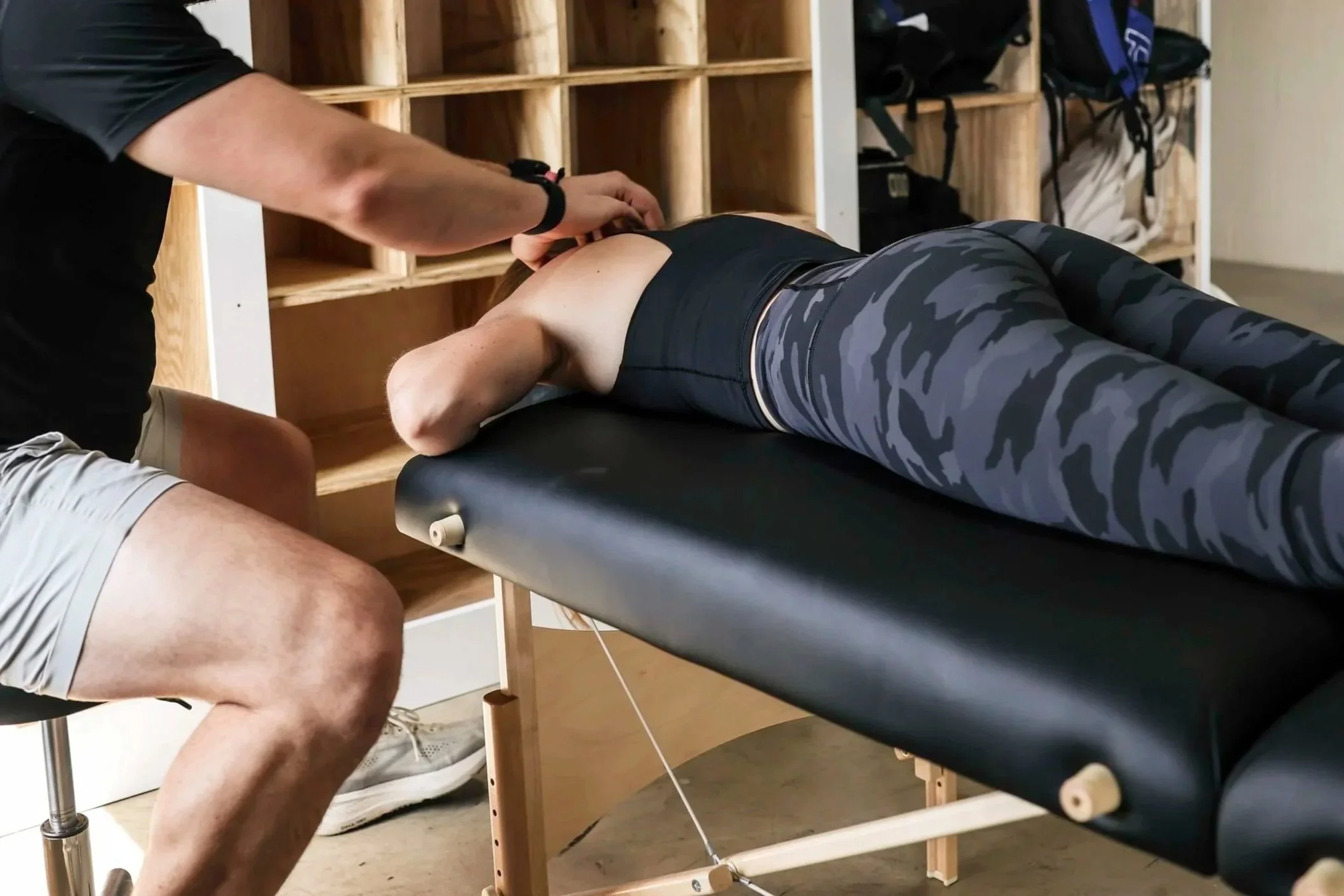 A person lying face down on a massage table receiving a back massage, with wooden shelves and backpacks visible in the background.