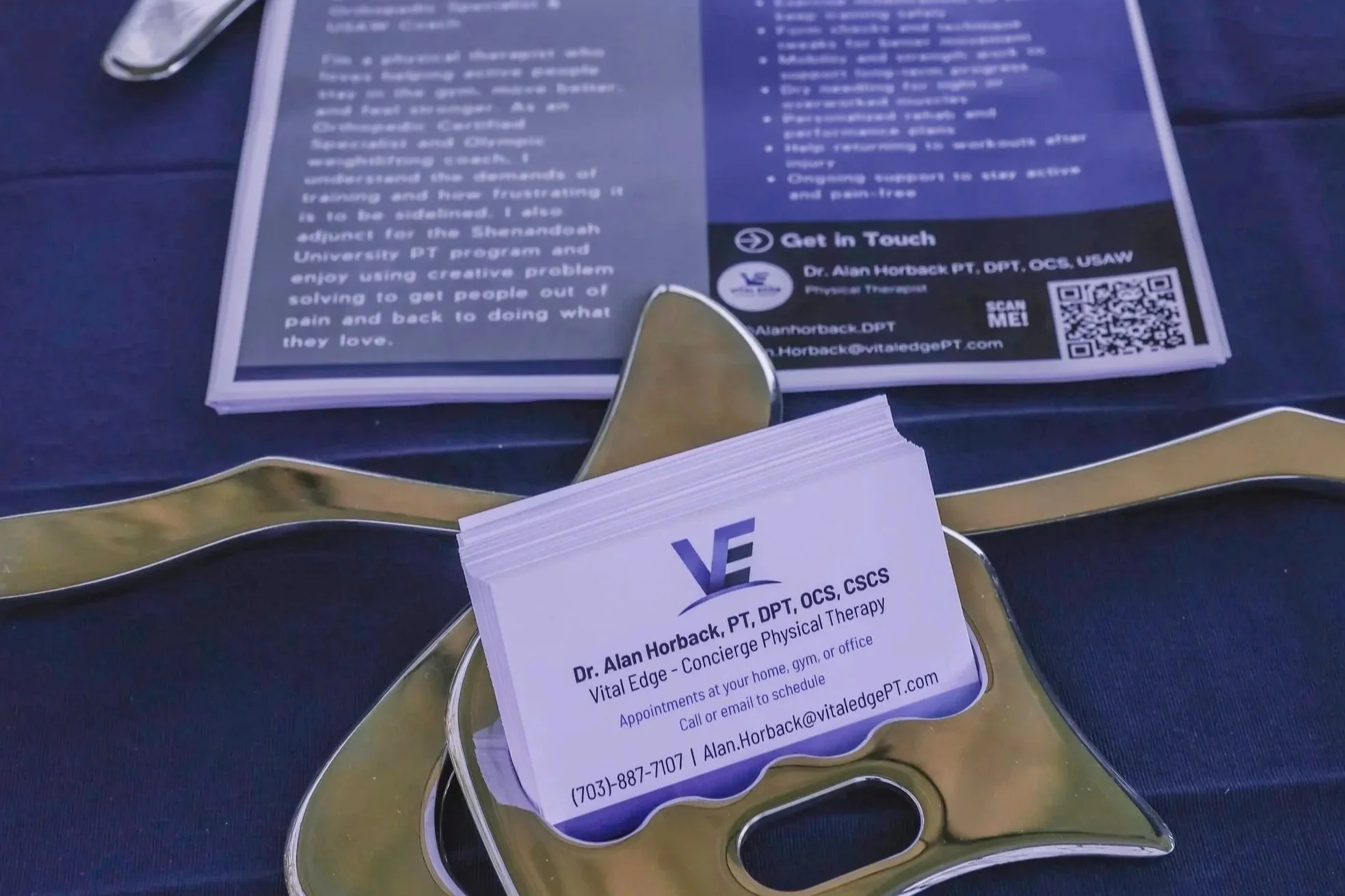 Close-up of a printed flyer or brochure placed on a reflective gold-colored tray, with a business card partially visible in the foreground. The flyer contains text and a QR code, mentioning Dr. Alan Horback, a physical therapist, along with contact information and a brief description of services offered.
