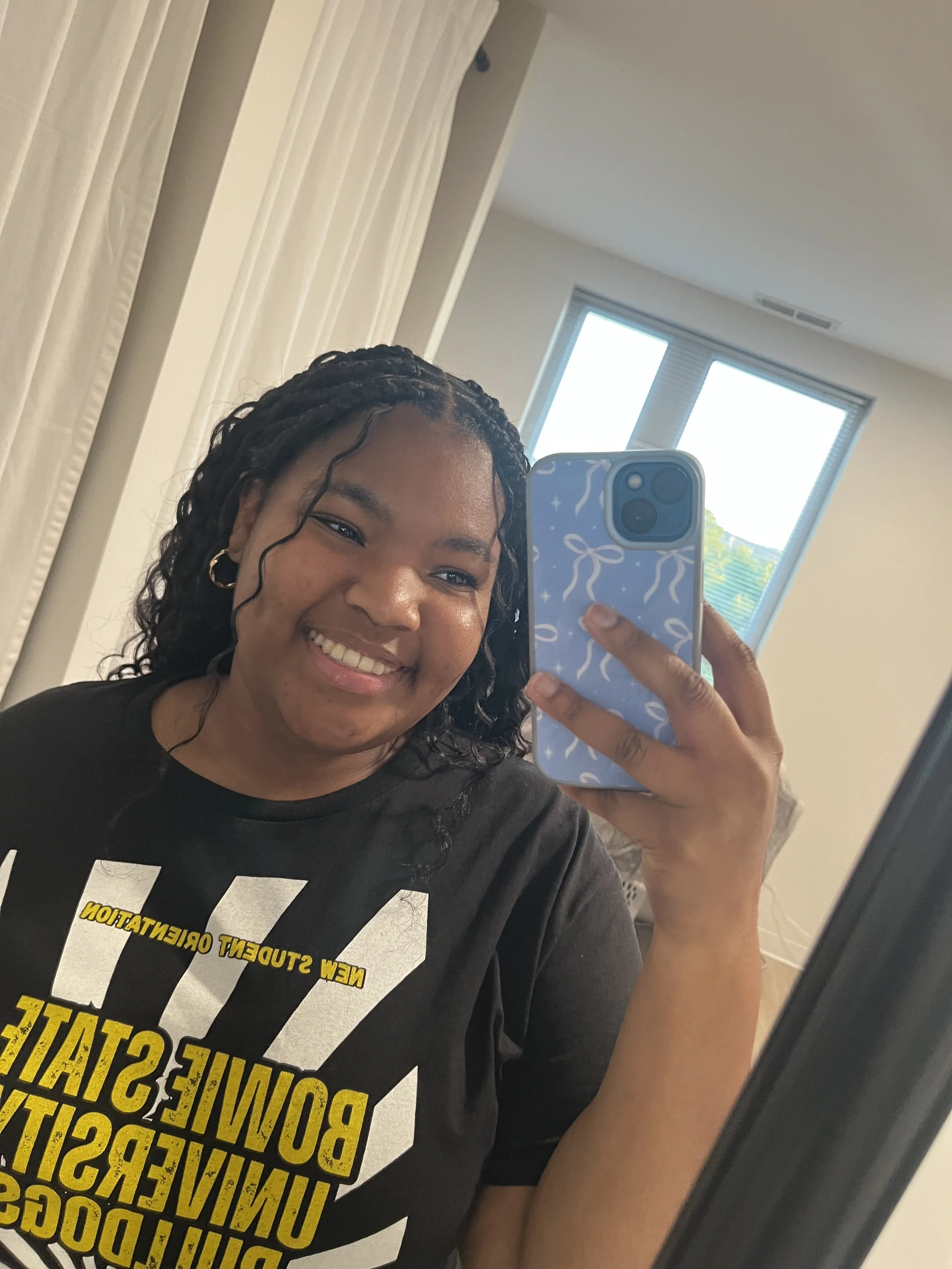 A young woman with curly hair taking a mirror selfie in a room with a window in the background. She is smiling and holding a blue phone with a patterned case.
