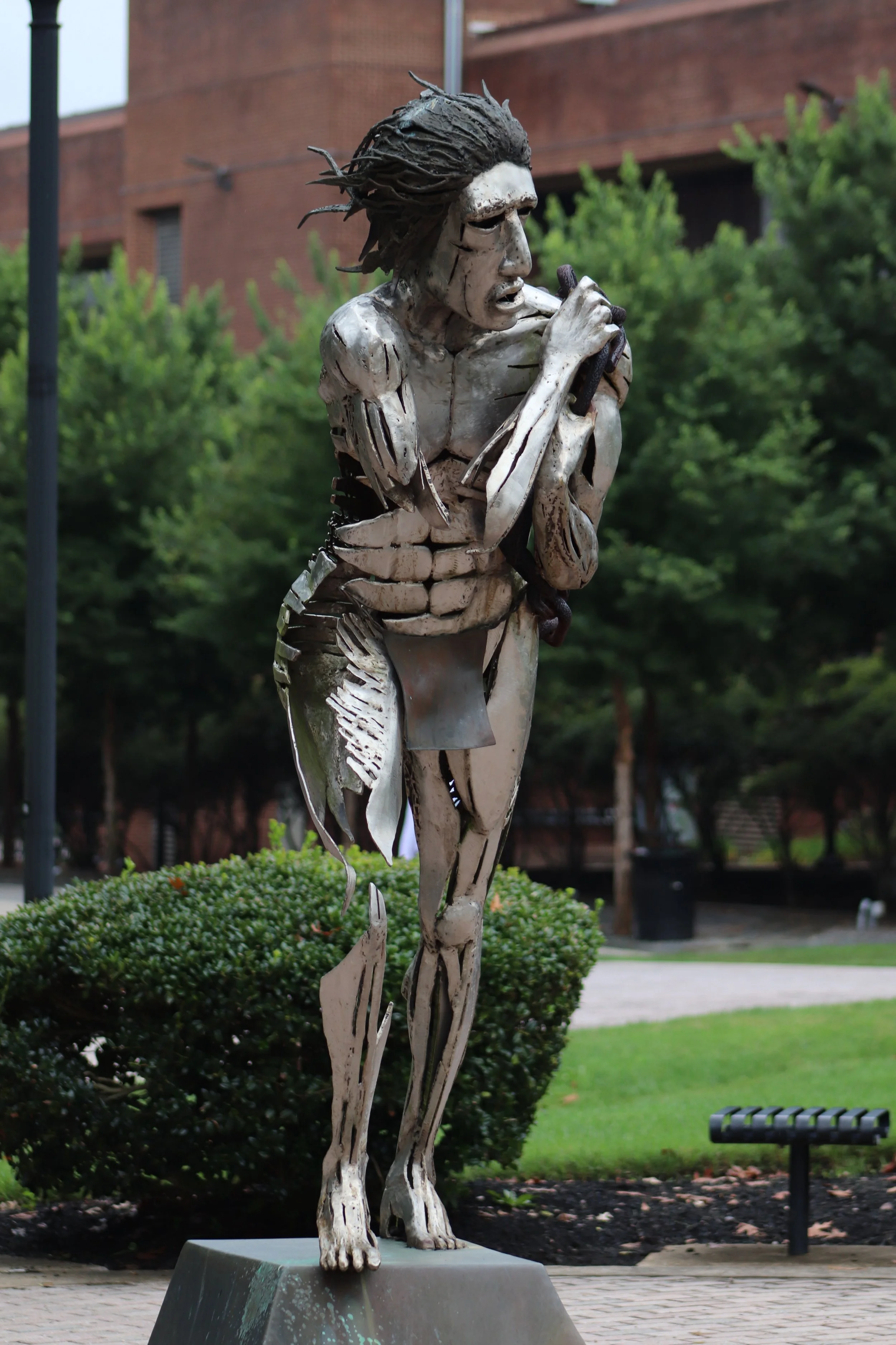 A metallic sculpture of a human figure playing a flute outdoors with trees and a brick building in the background.